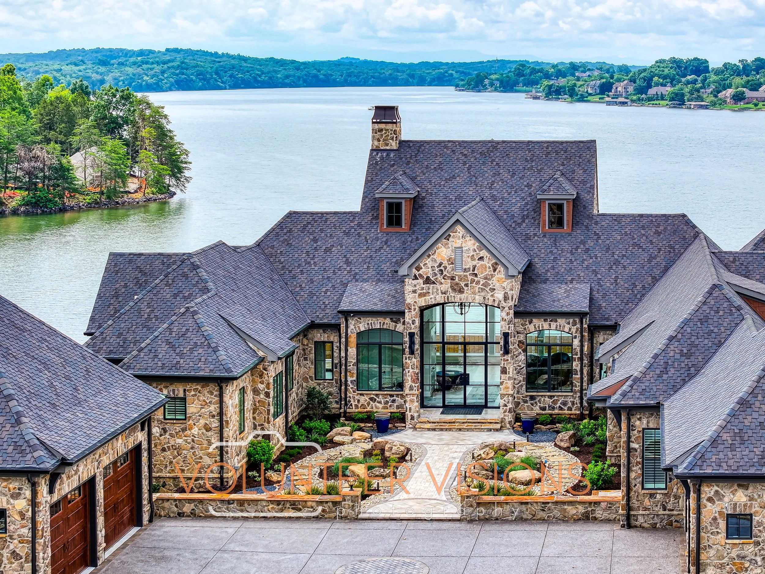 Luxury stone and brick house with large windows, gray shingle roof, landscaped garden, and a view of a lake with trees and houses across the water.