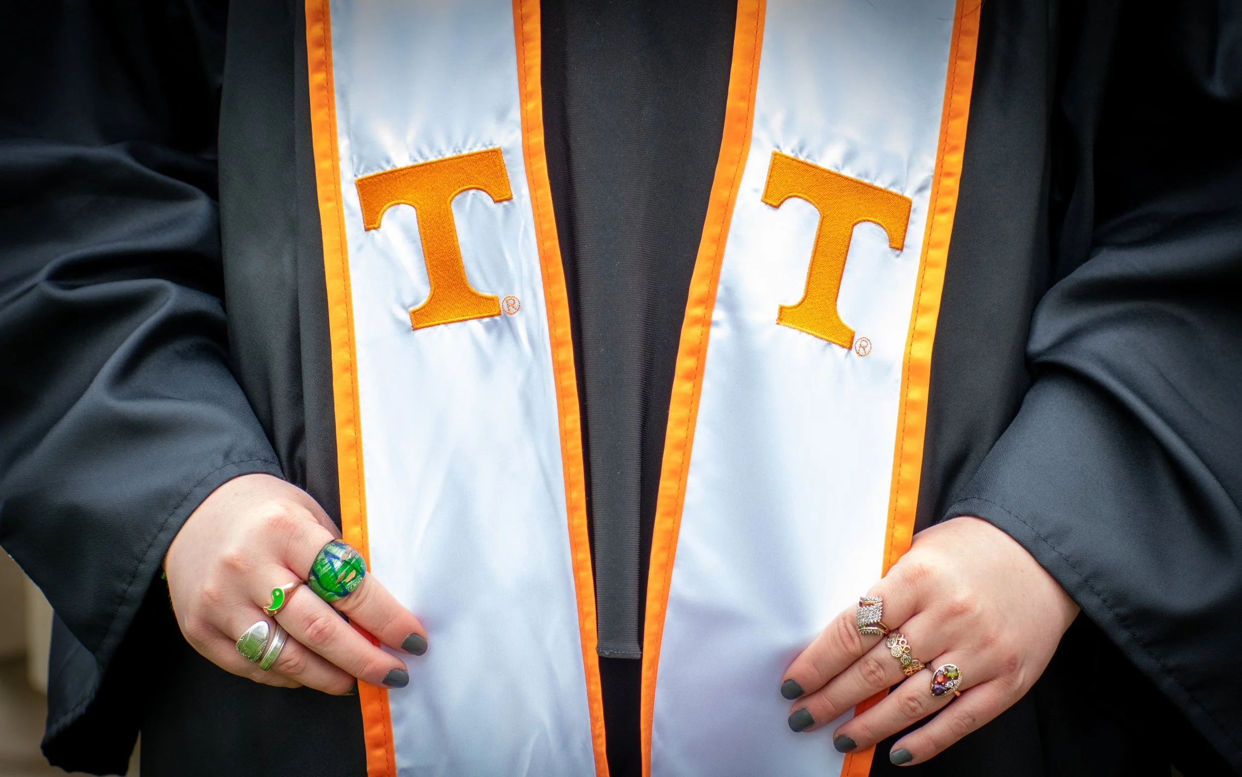 Close-up of a person wearing a black graduation gown and a white stole with orange border and the University of Tennessee logo, featuring large orange embroidered letters 'T'. The person is holding their hands in front, showing rings and painted nail