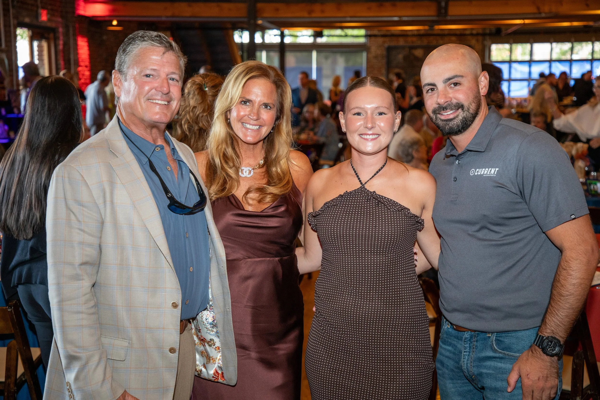 Four people standing closely together and smiling at an indoor event. Background shows a large crowd and a rustic interior with brick walls and large windows.