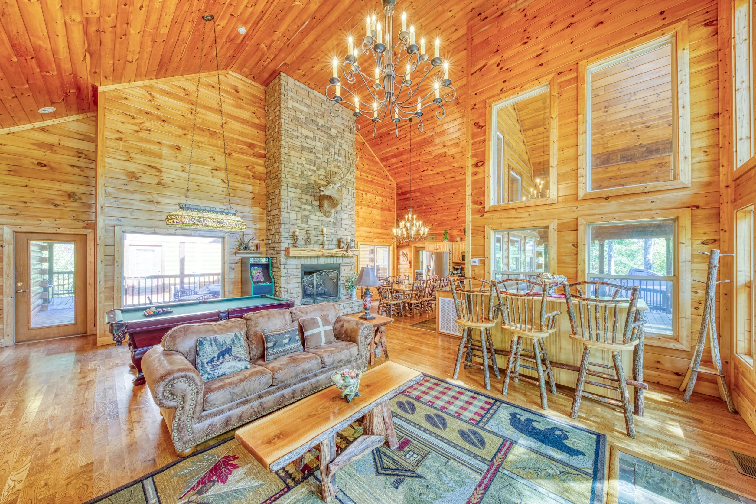 A spacious living room in a log cabin with high wooden ceilings and large windows. The room features a brick fireplace with a mounted deer head above it. There is a leather sofa with decorative pillows, a wooden coffee table, a colorful area rug with
