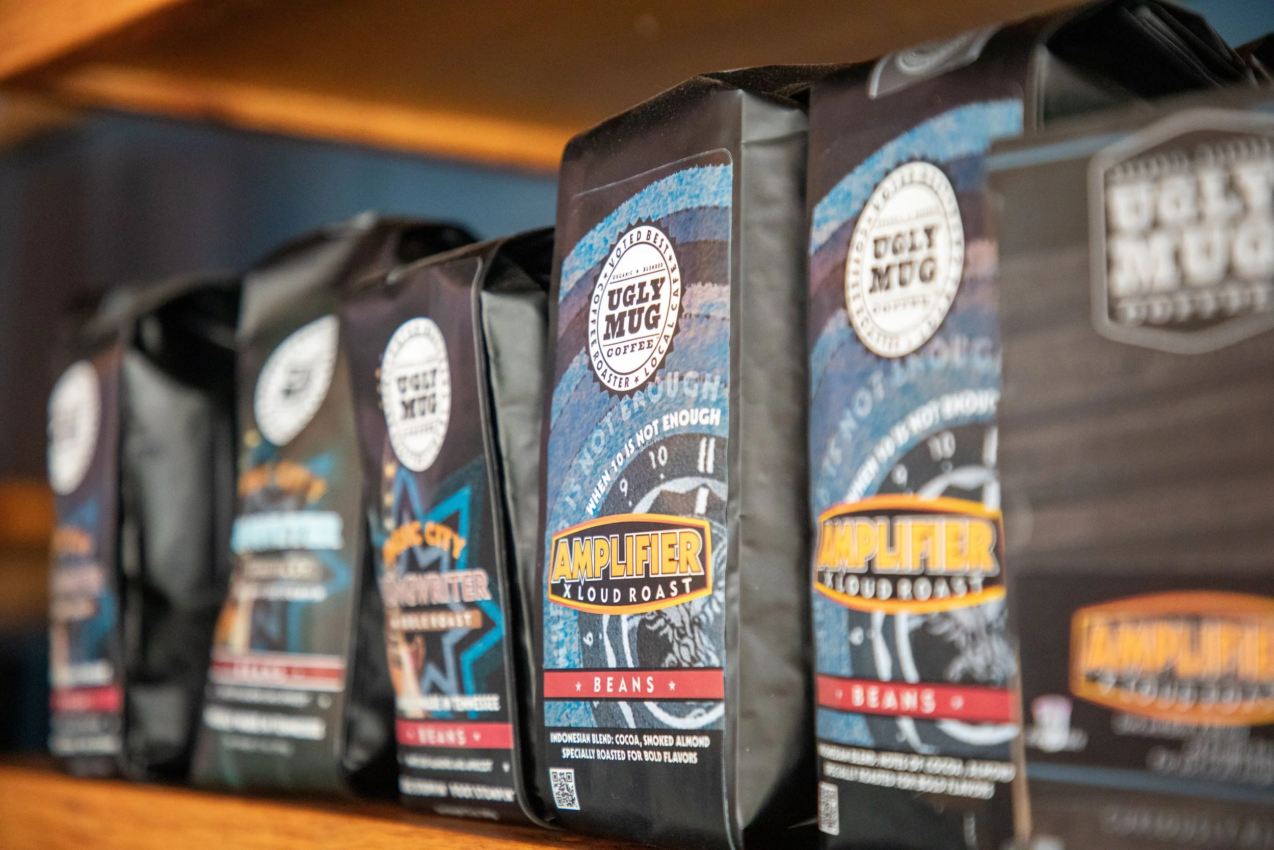 Rows of Ugly Mug coffee bags on a wooden shelf, labeled as 'AMPLIFIER XLoud Roast Beans' with a colorful design.
