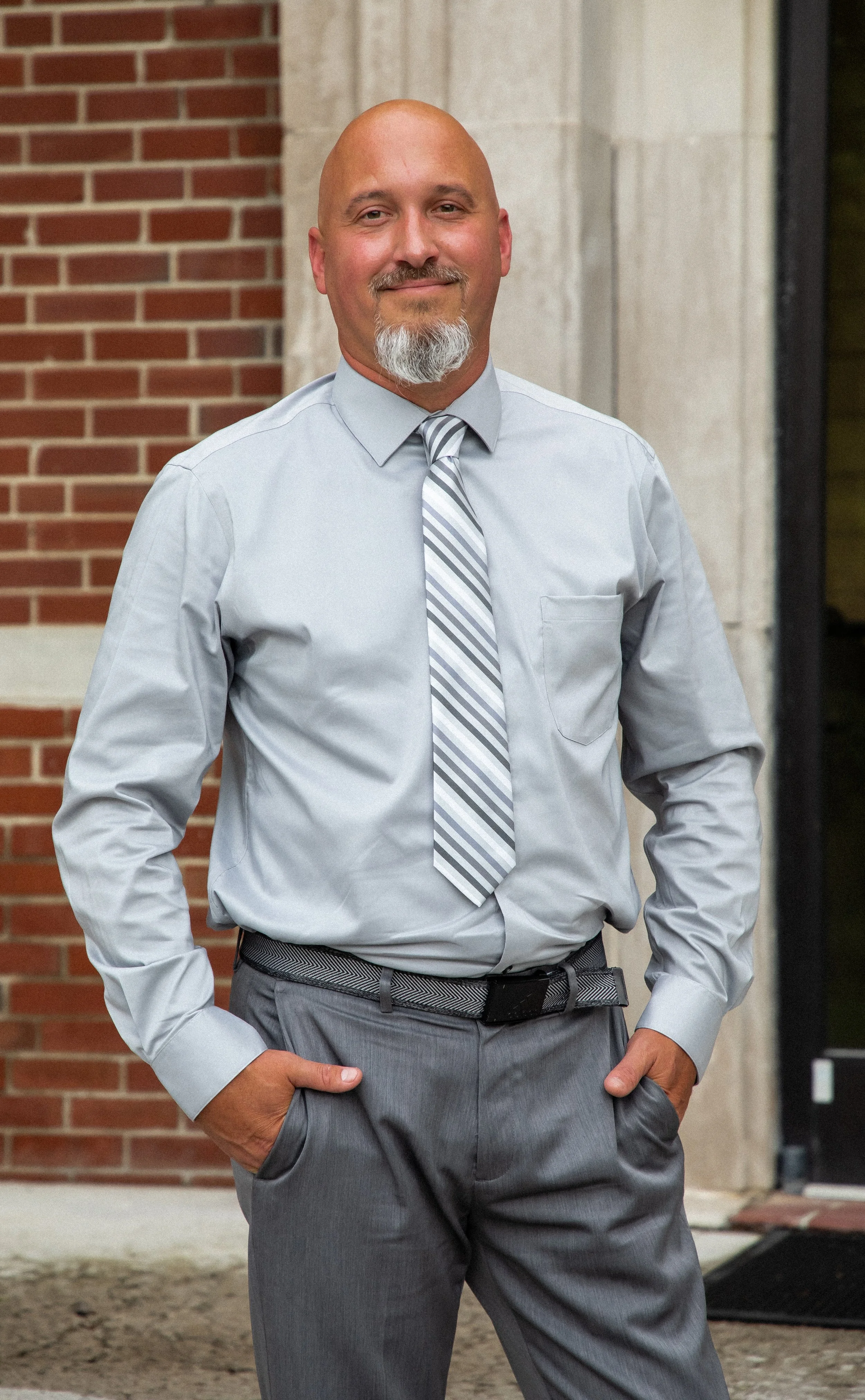 A middle-aged man with a bald head and a gray goatee smiling while standing outside in front of a brick wall, wearing a light gray dress shirt, striped tie, and gray dress pants.