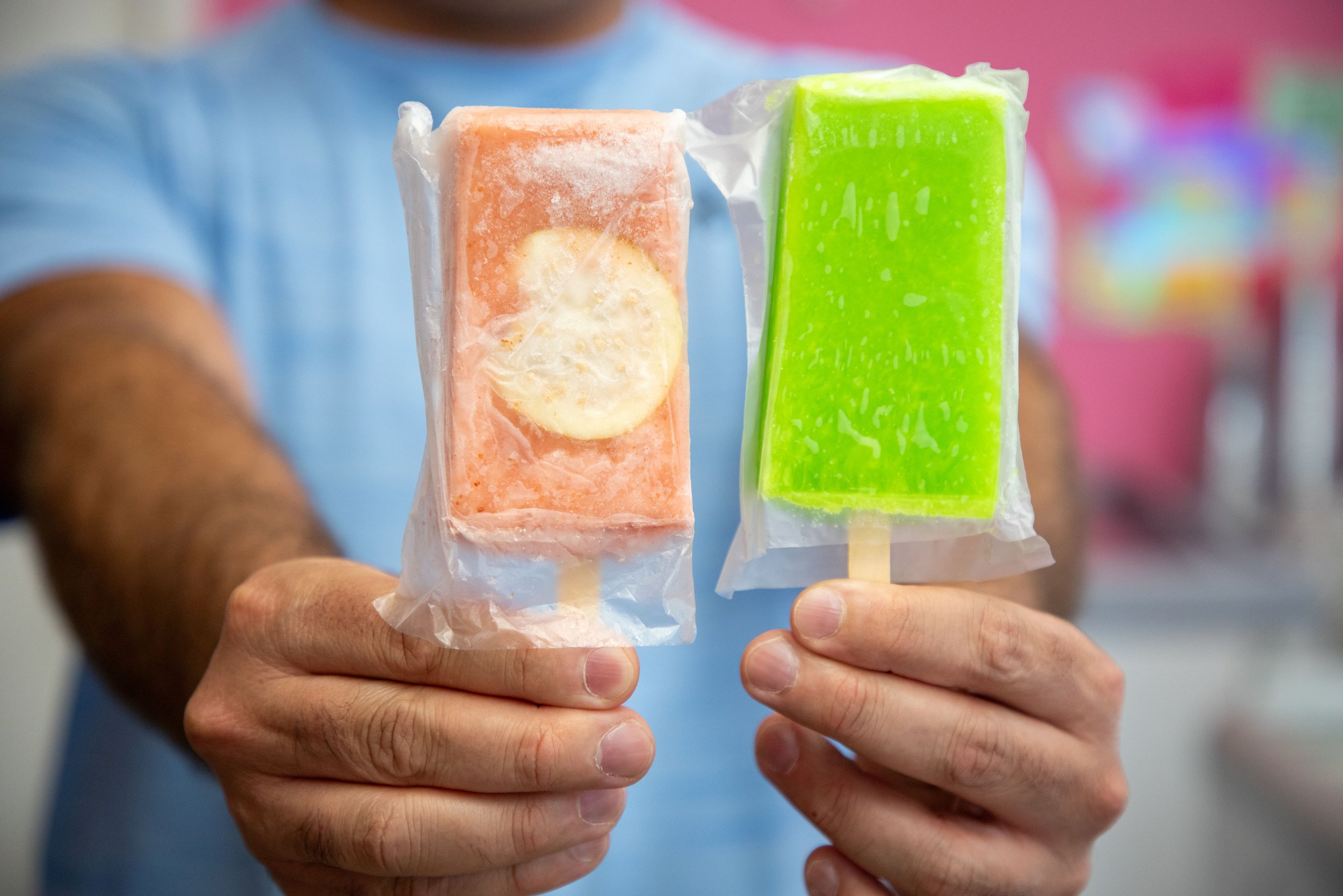 A person holding two popsicles, one orange and one bright green, both in clear plastic wrappers, with a blurred background.