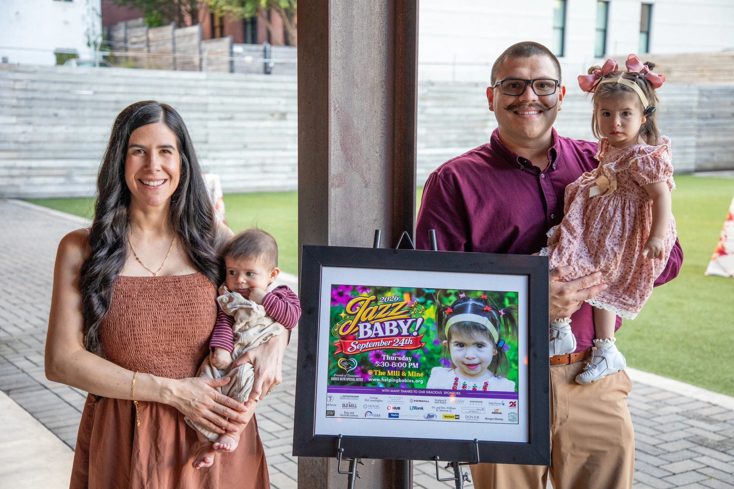 A woman holding a young child in her arms stands next to a man holding a young girl. They are outdoors near a poster advertising a jazz baby event. The woman has long dark hair and wears a brown dress, the girl has pigtails with pink bows, the man ha