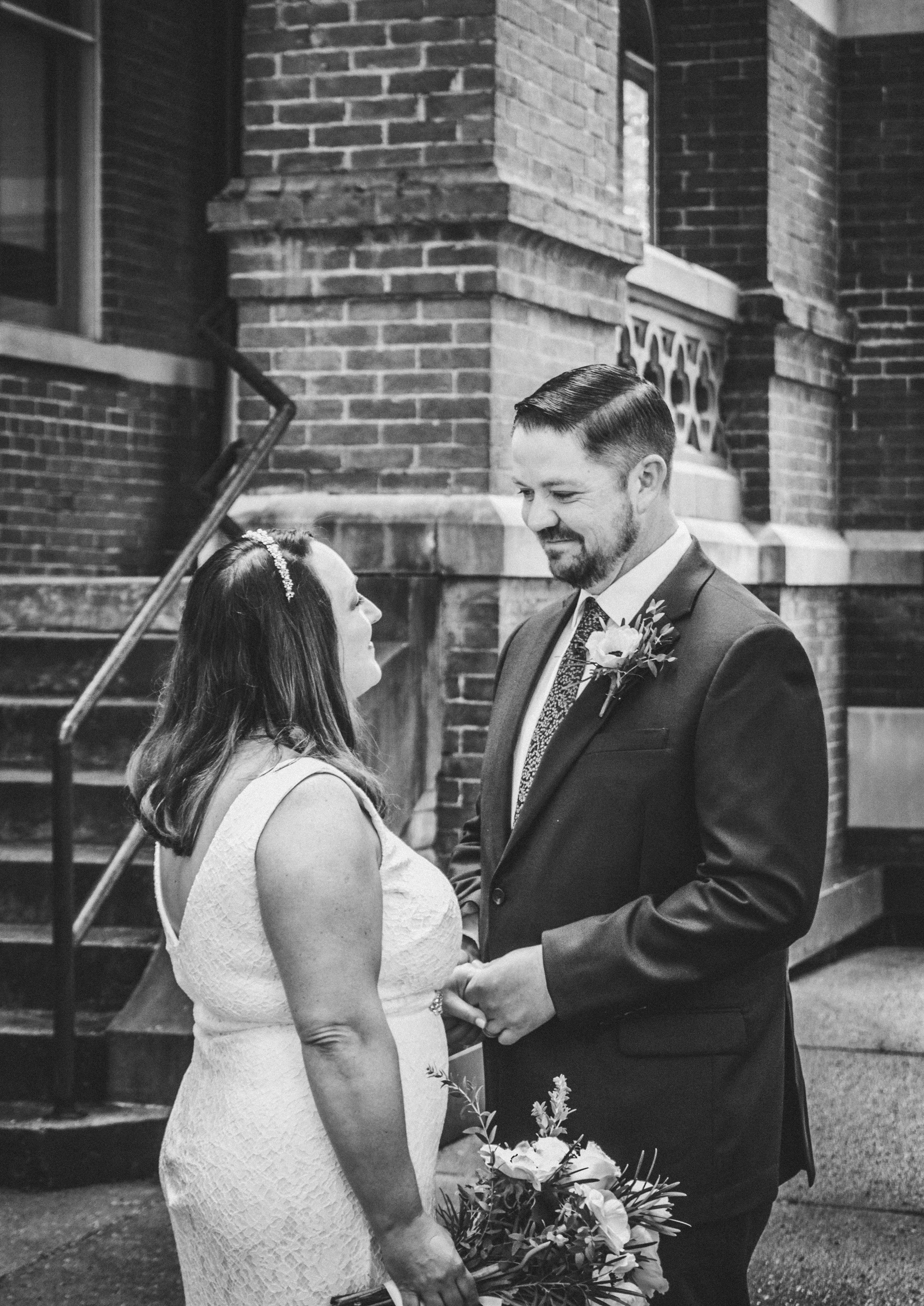 A black and white photo of a bride and groom standing outdoors in front of a brick building, holding hands and gazing at each other during their wedding ceremony. The bride wears a white lace dress with a headband, and the groom is in a dark suit wit