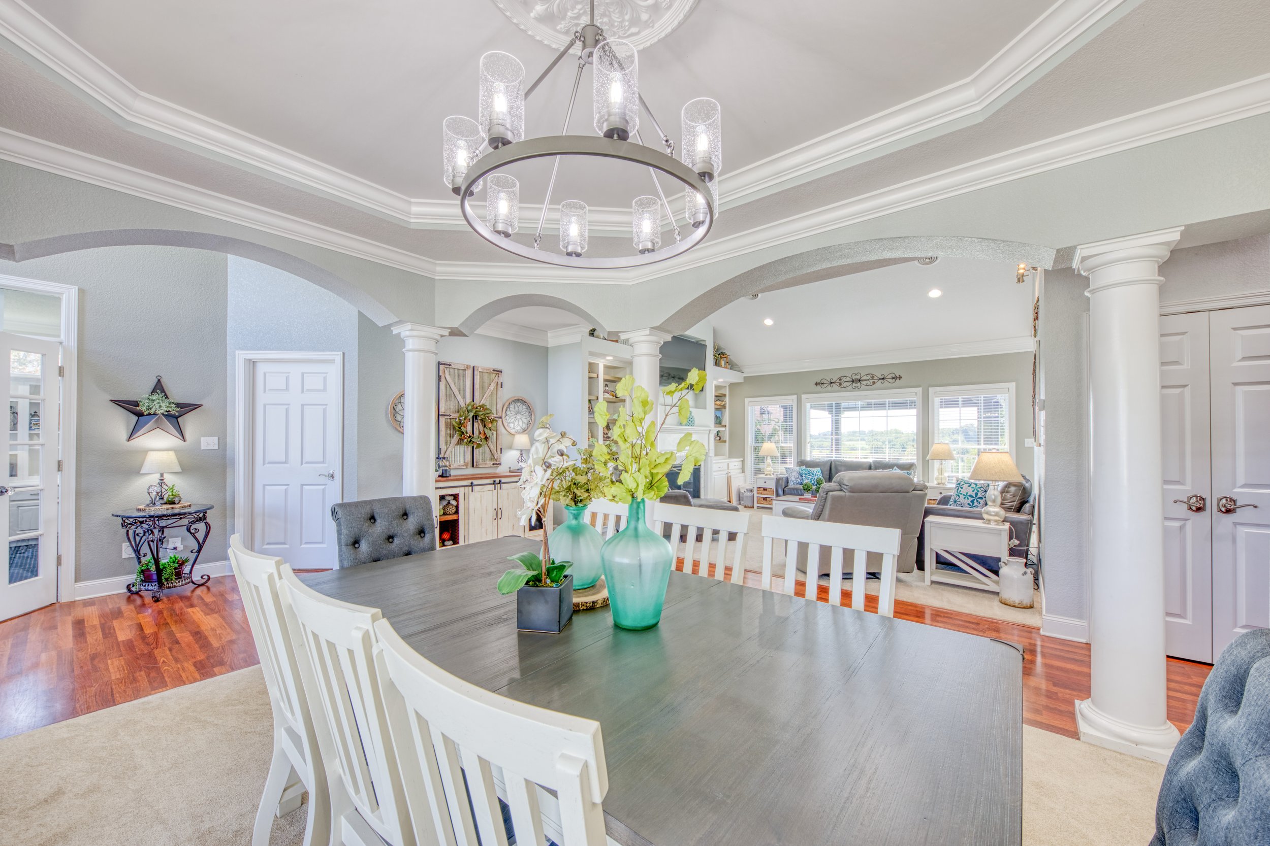A spacious dining area with a large wooden table surrounded by white chairs. On the table, there are three green vases with plants. The room features crown molding and a modern chandelier with multiple glass cylindrical shades hanging from the ceilin