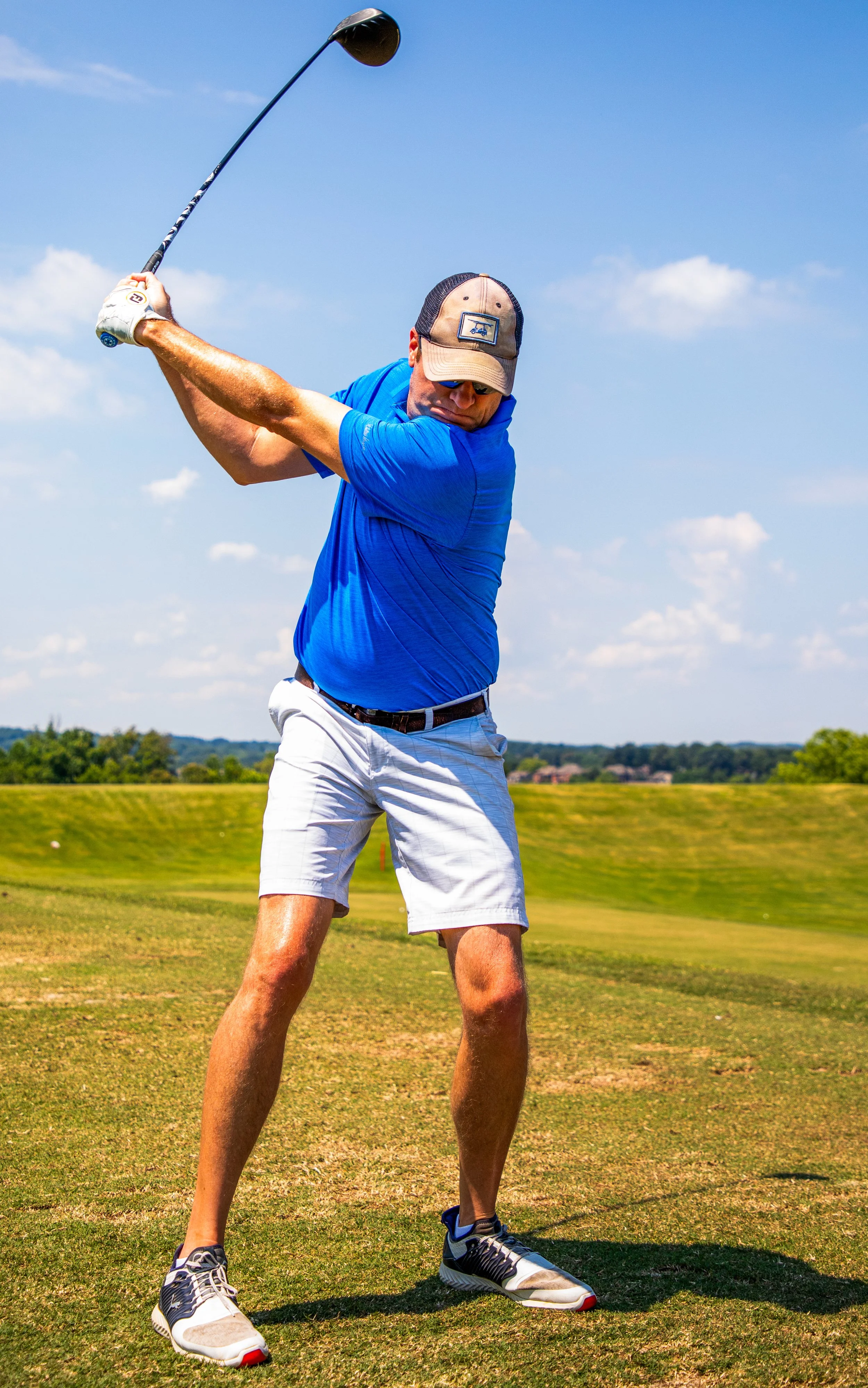 A man wearing a blue polo shirt, gray shorts, and a beige cap playing golf on a sunny day, swinging a golf club on a grassy course with a blue sky and scattered clouds.