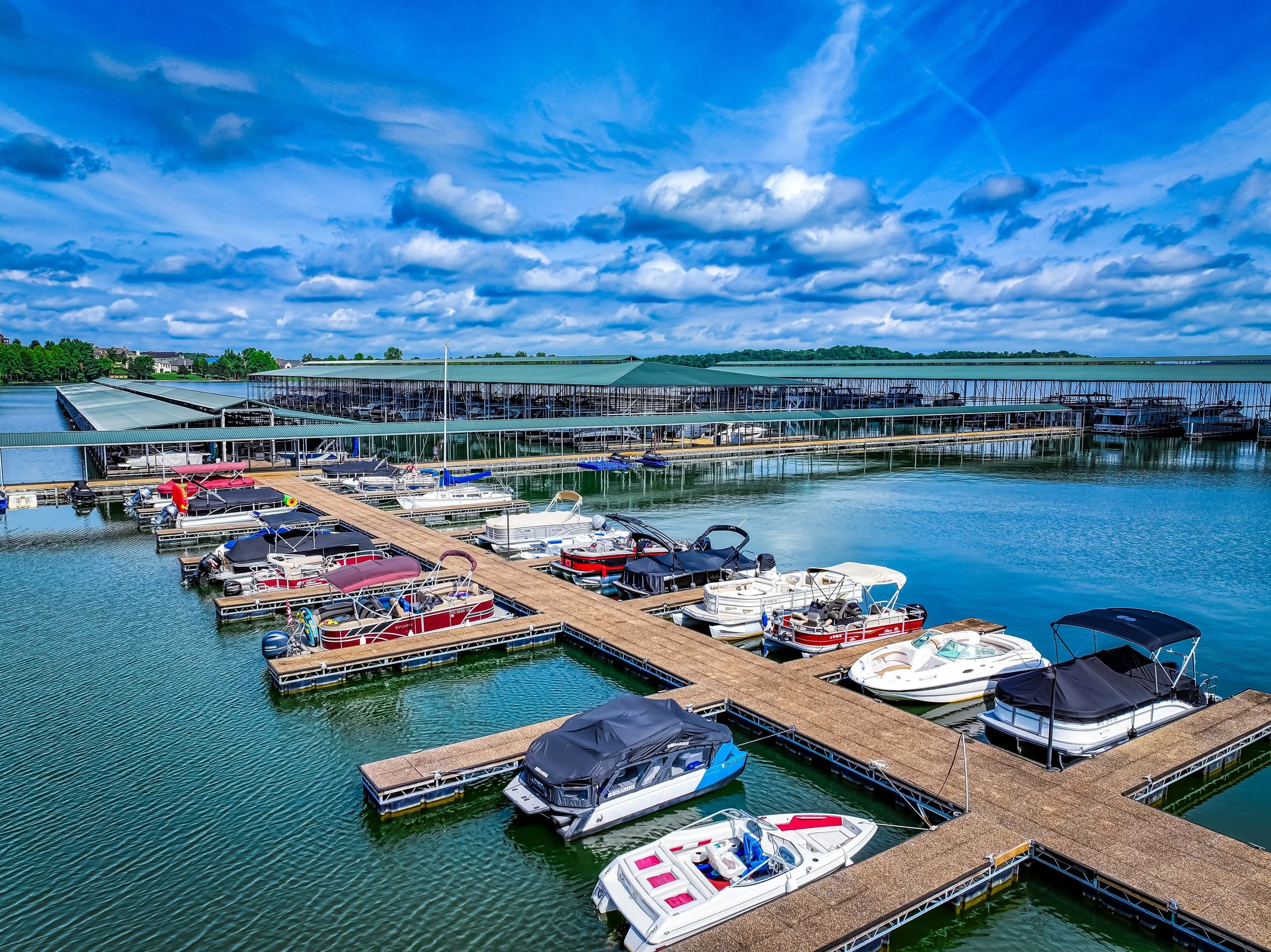 View of a marina with multiple boats docked along floating docks under a partly cloudy blue sky, with a bridge and tree-covered hills in the background.