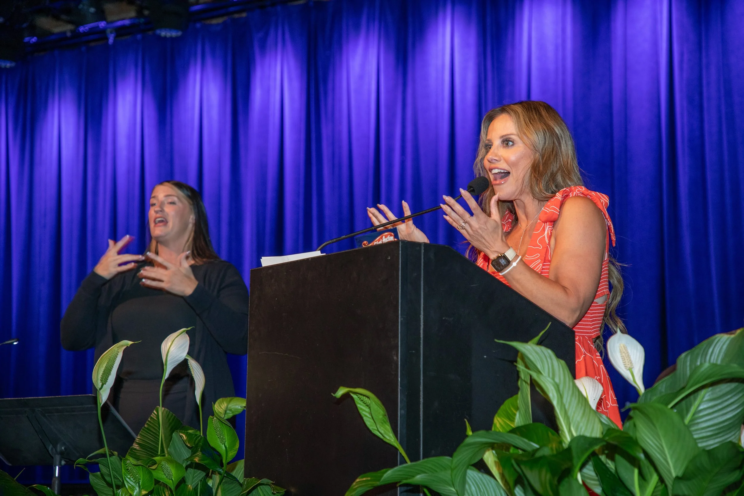 Two women speaking at a podium on stage with blue curtains. One woman is wearing a red and white patterned dress, and the other is dressed in black, using sign language. Green plants are in the foreground.