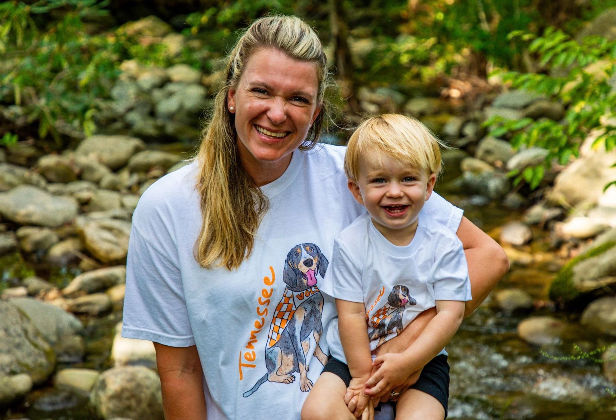 A woman and a young boy smiling outdoors near a stream with rocks and green foliage in the background. They are wearing white shirts with dog illustrations.