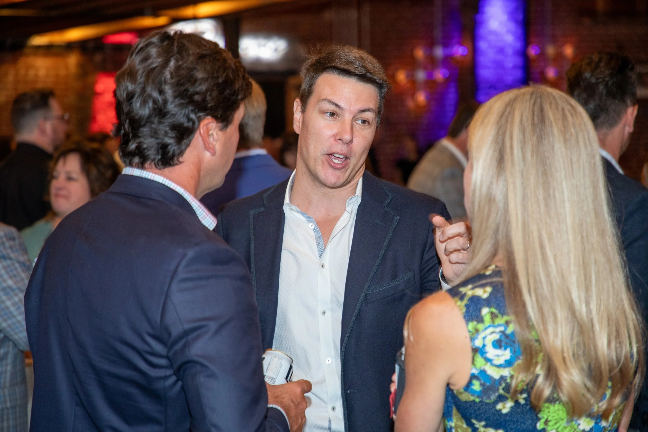 Three people engaged in a conversation at an indoor event, with a brick wall and colorful lighting in the background.
