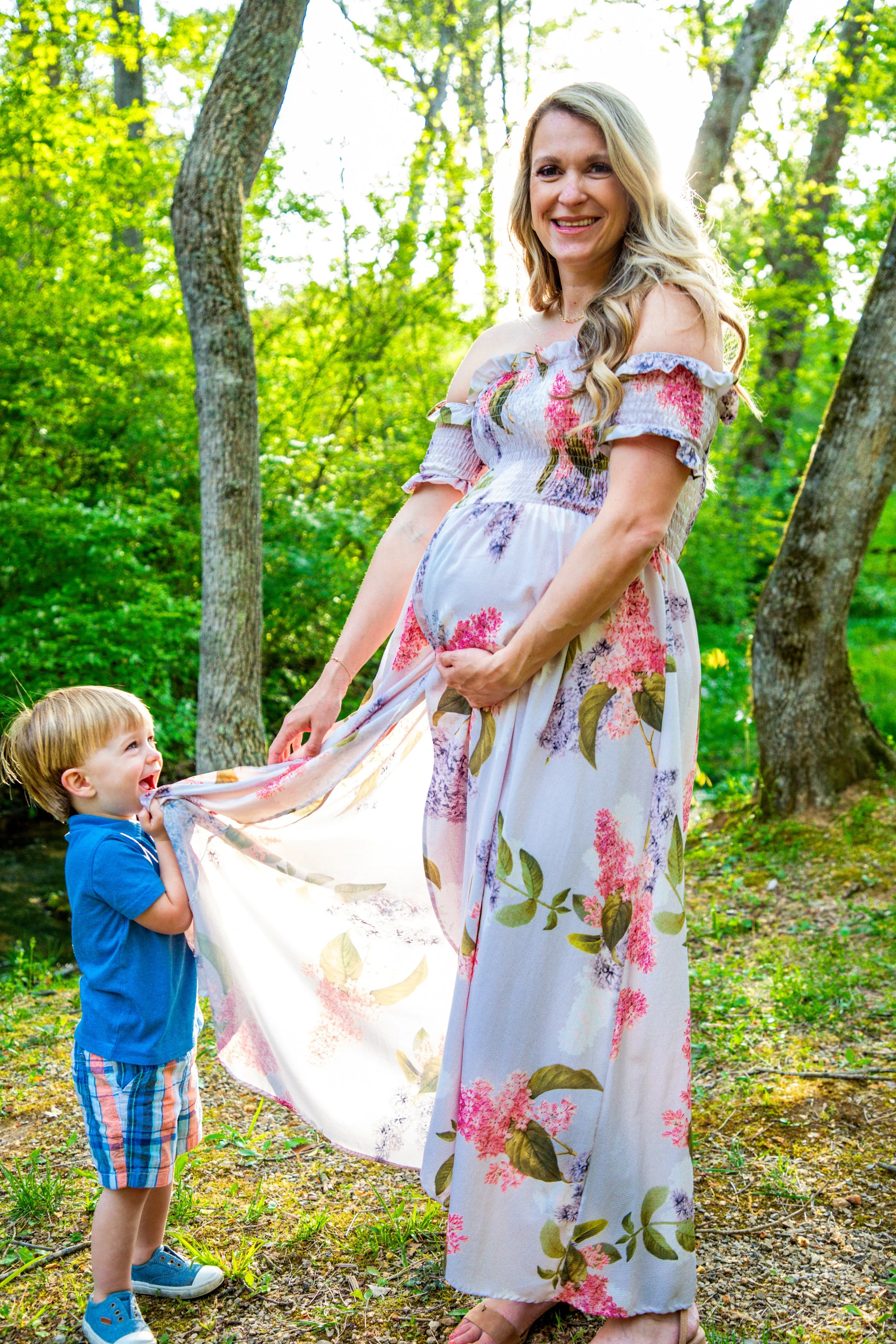 A pregnant woman in a floral dress smiling outdoors in a forested area, with a young boy pulling the dress and looking up at her, both enjoying a playful moment.