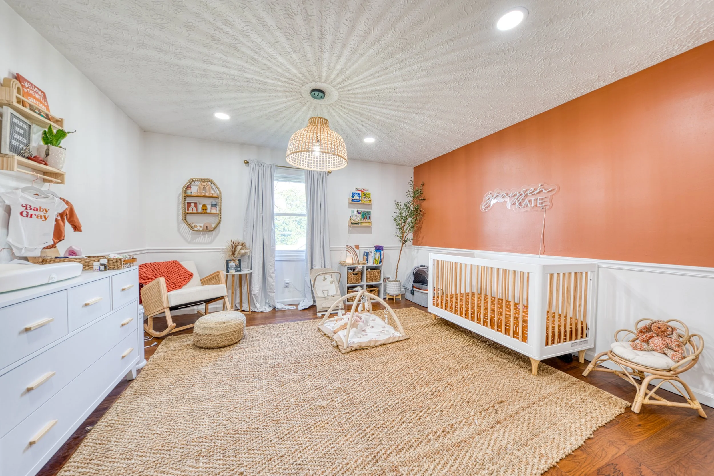 Nursery room with white dresser, wicker rocking chair, changing table, crib, shelf with toys, and framed decor, illuminated by ceiling lights and a hanging paper pendant