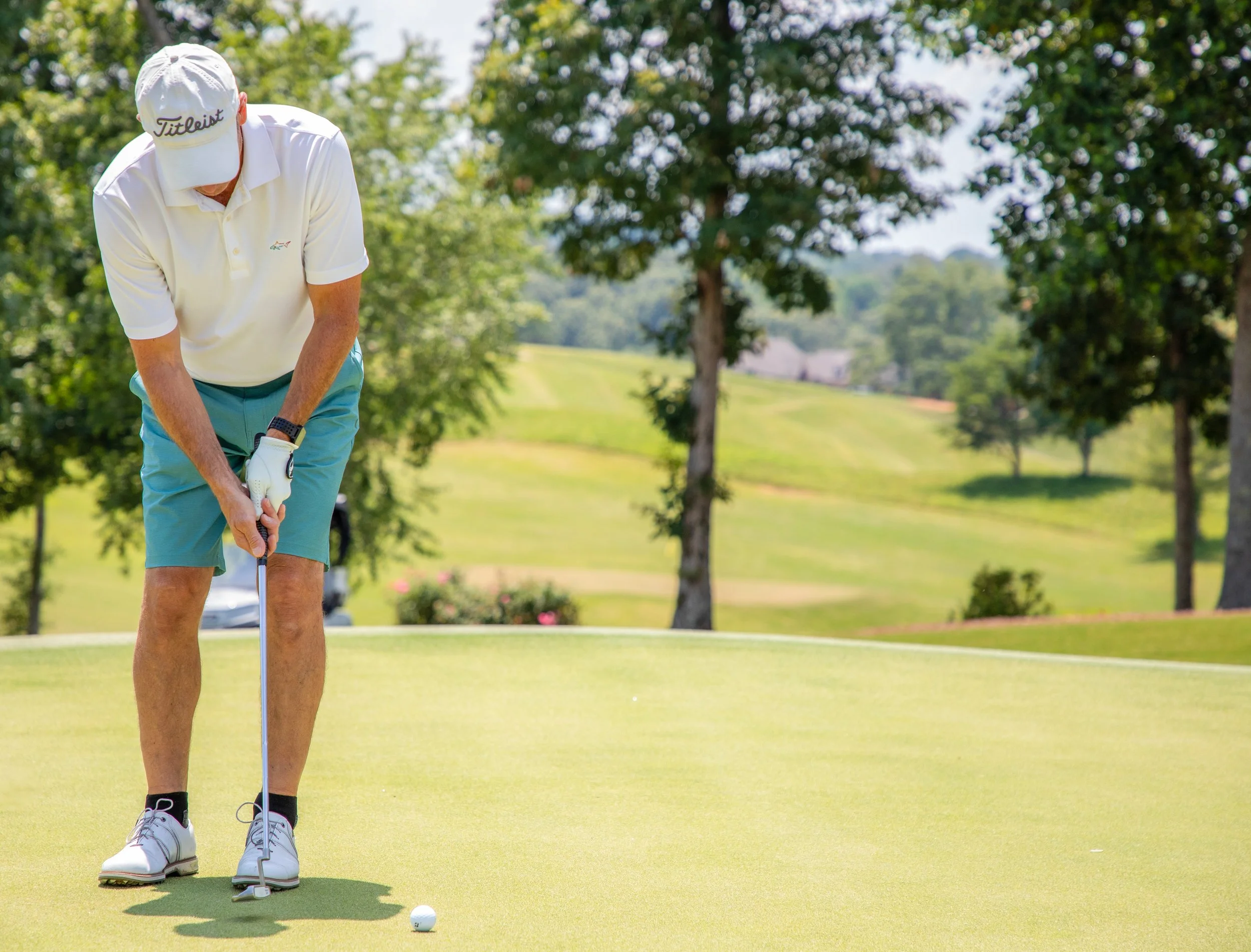 A man wearing a white golf shirt, shorts, and a white cap with the Titleist logo practicing putting on a golf course, with trees in the background.