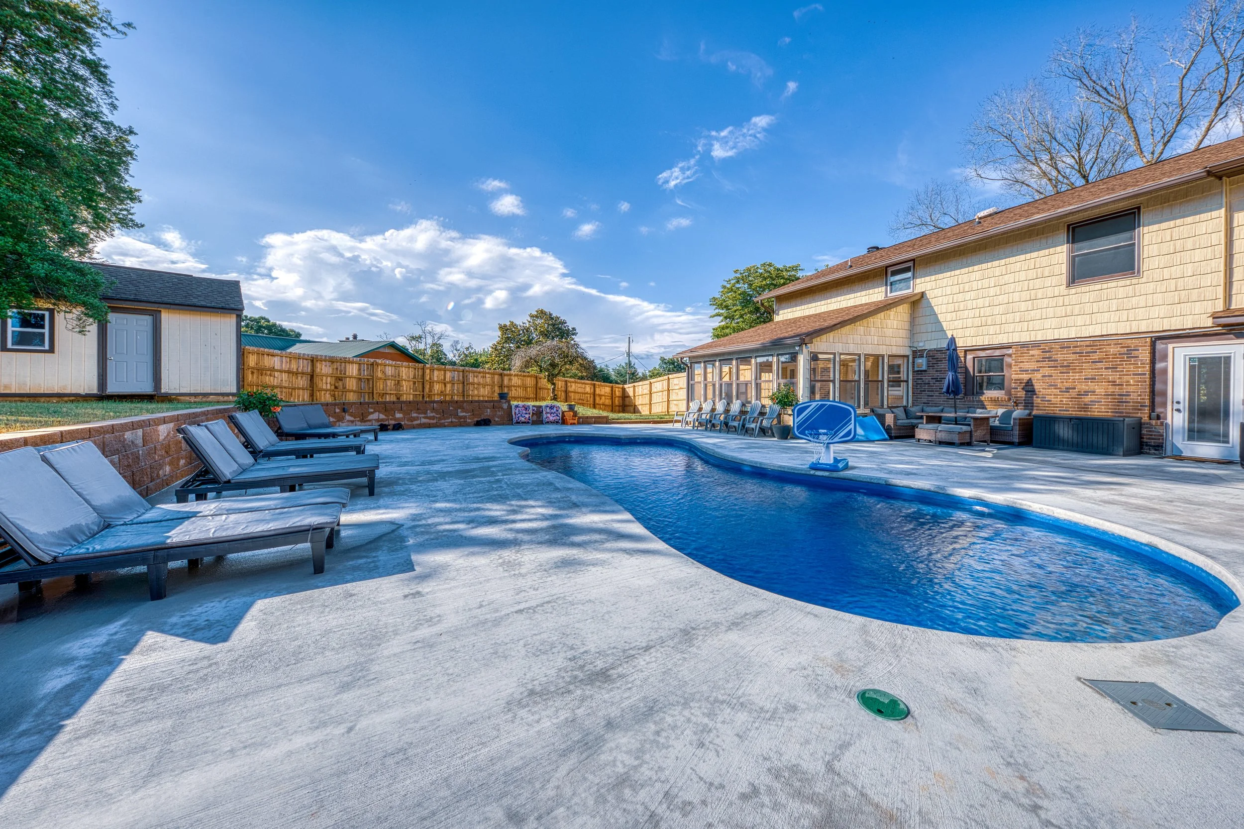 Backyard with a swimming pool, lounge chairs, patio furniture, basketball hoop, and a sunny sky.