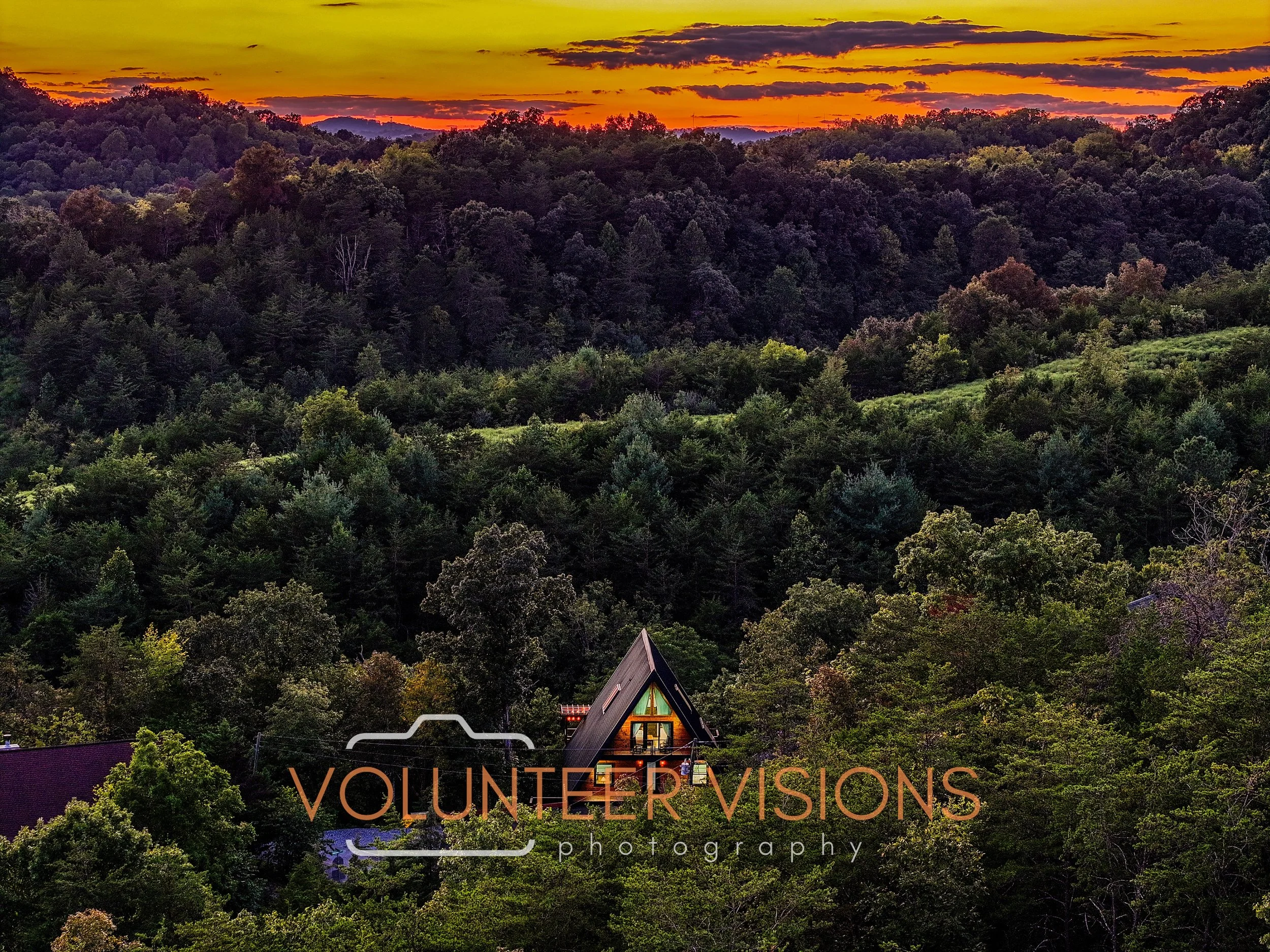 A house with lights on, situated among a dense forest of green trees, with a colorful sunset sky overhead.