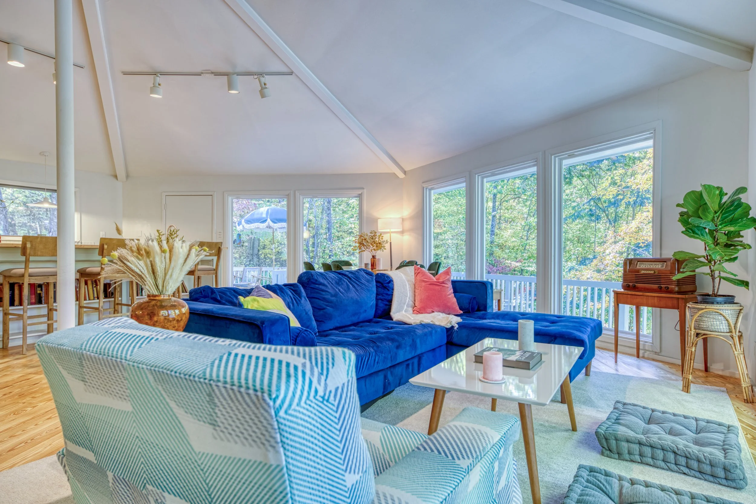 Living room with blue velvet sofa, striped armchair, coffee table with candles, large windows showing trees outside, plants, and a dining area with wooden chairs and a bar counter.