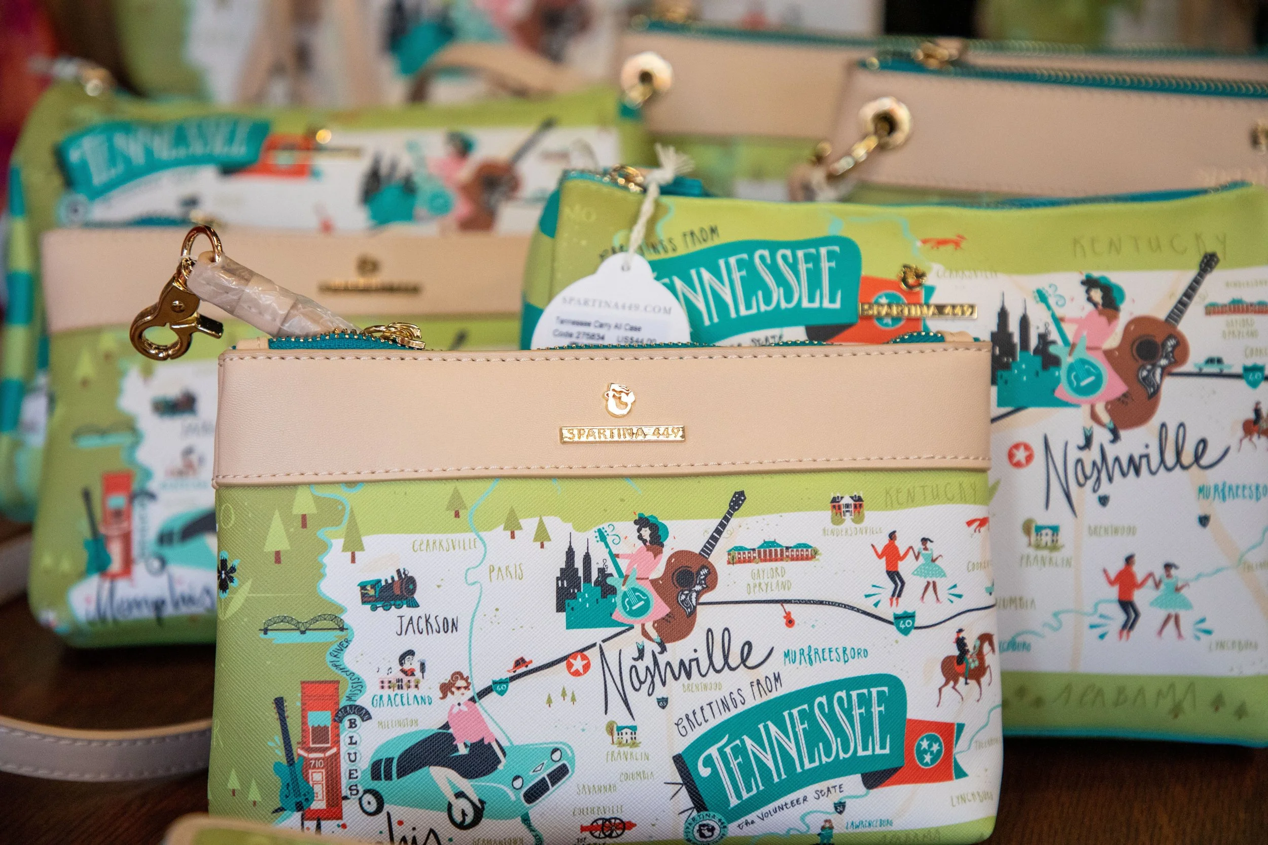 Multiple small pouches with colorful Tennessee-themed maps on them, featuring illustrations of landmarks, people, and animals, with gold zipper pulls and tags.