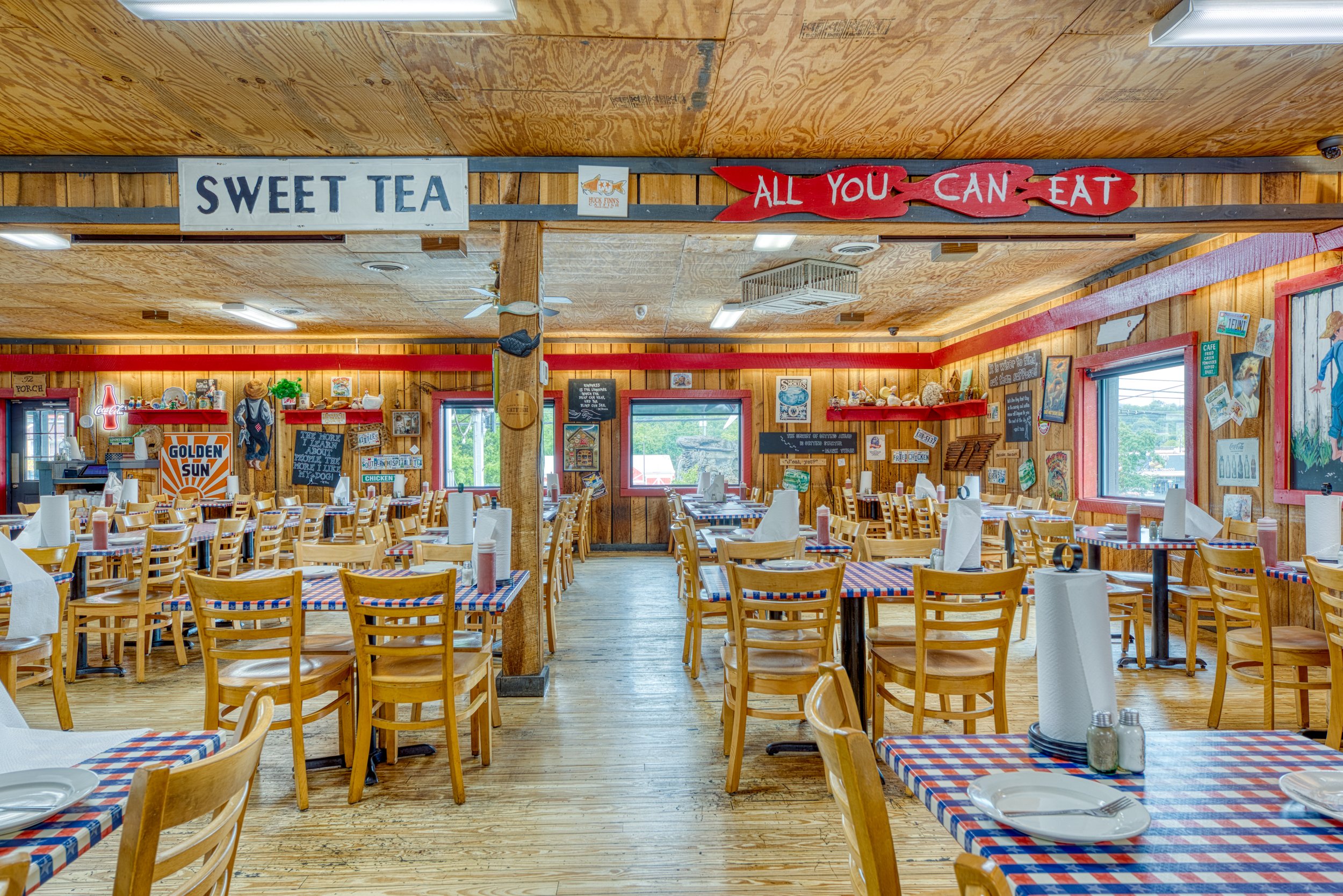 A rustic restaurant interior with wooden walls and matching wooden chairs with checkered red, white, and blue tablecloths, set with plates, utensils, and napkins. Decorative signs and artwork adorn the walls.