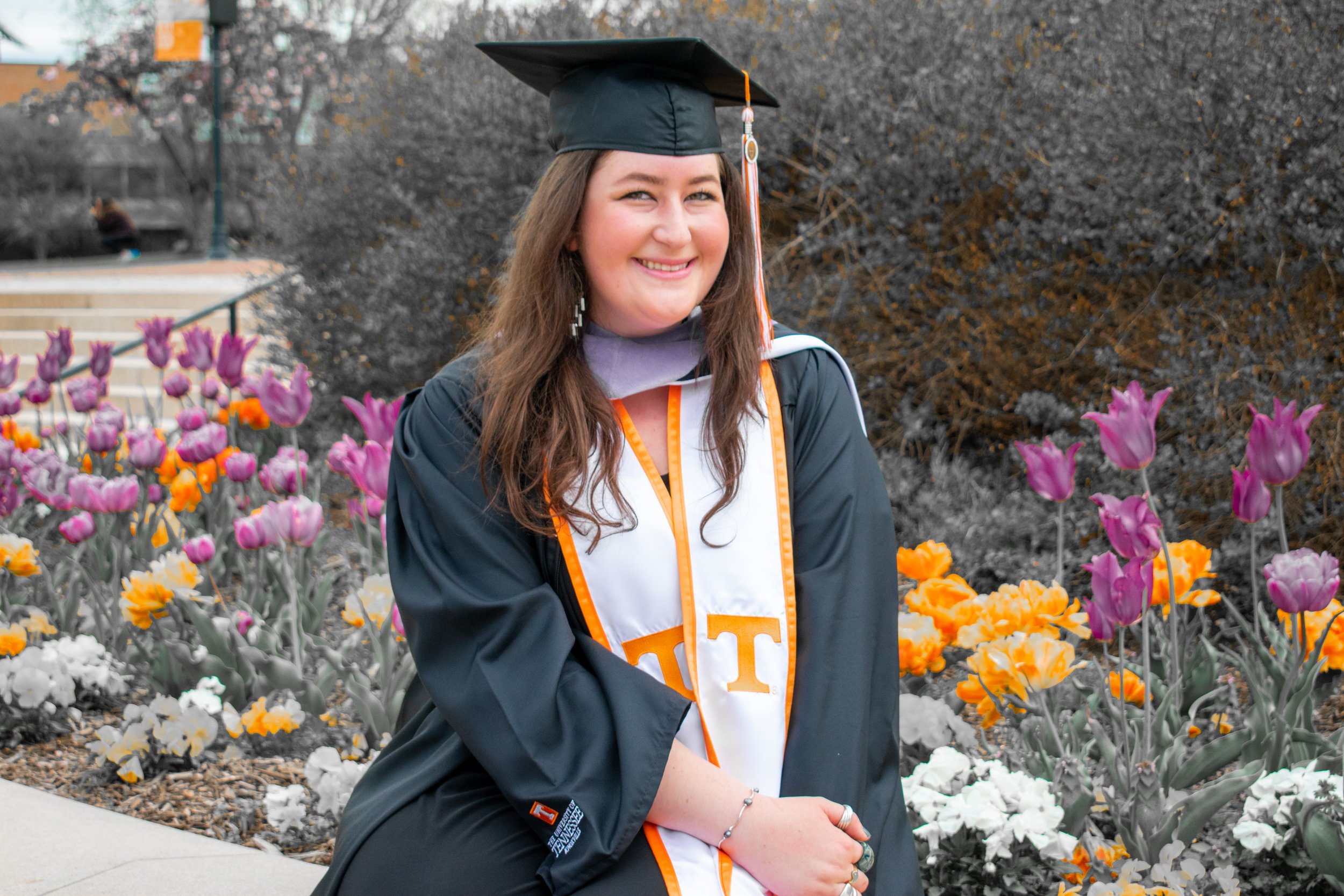 A young woman in a graduation cap and gown seated outdoors in front of flowers, smiling at the camera.