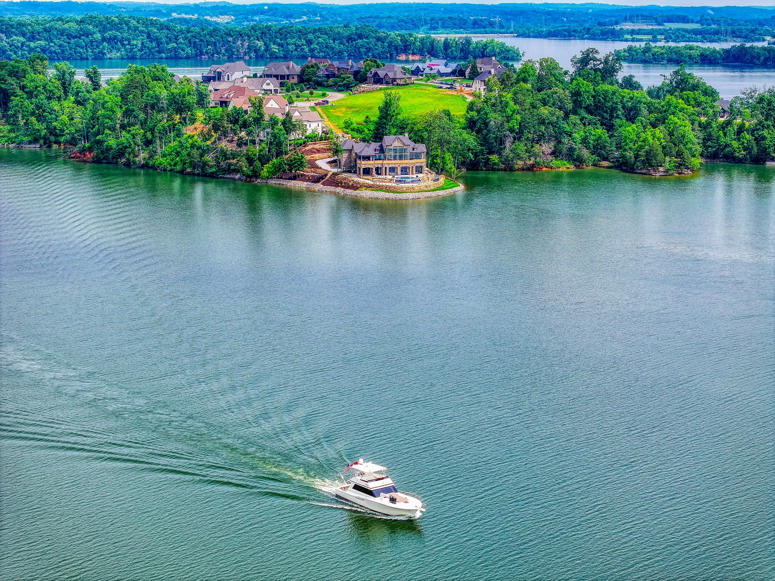 A white motorboat with a red, white, and blue flag on the stern moving across a calm lake with a visible wake behind it. The shoreline features a large house with a stone foundation and multiple decks, surrounded by lush green trees. In the backgroun