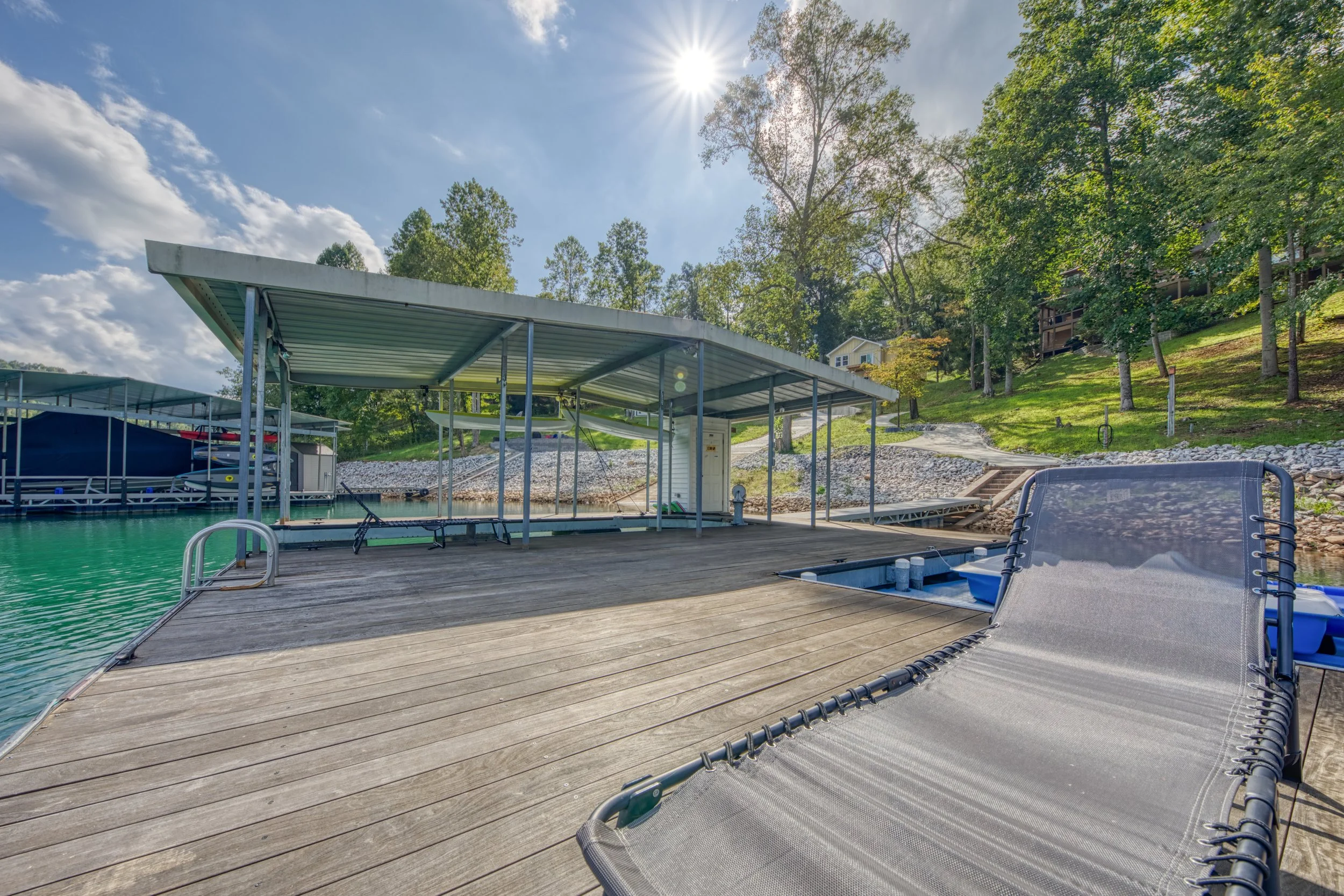 A lakeside dock with a lounge chair, surrounded by trees, with a metal covered boat slip and green water under a blue sky with sunlight.