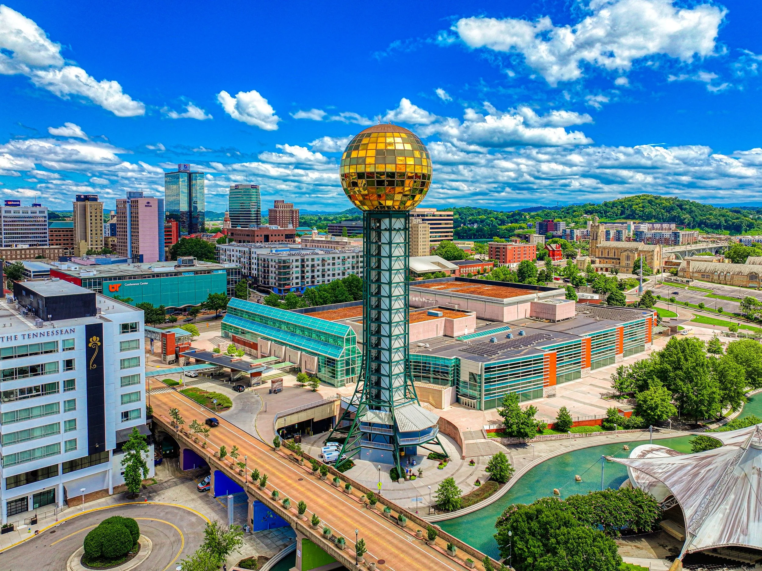 City skyline with tall modern buildings, hills with green trees in the background, and a prominent spherical golden structure on a tall pole in the foreground, under a blue sky with scattered clouds.