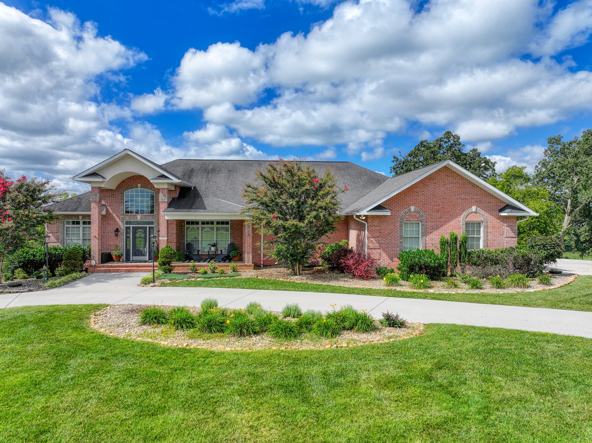 A brick house with a well-maintained front yard, trees, and a sidewalk under a partly cloudy blue sky.
