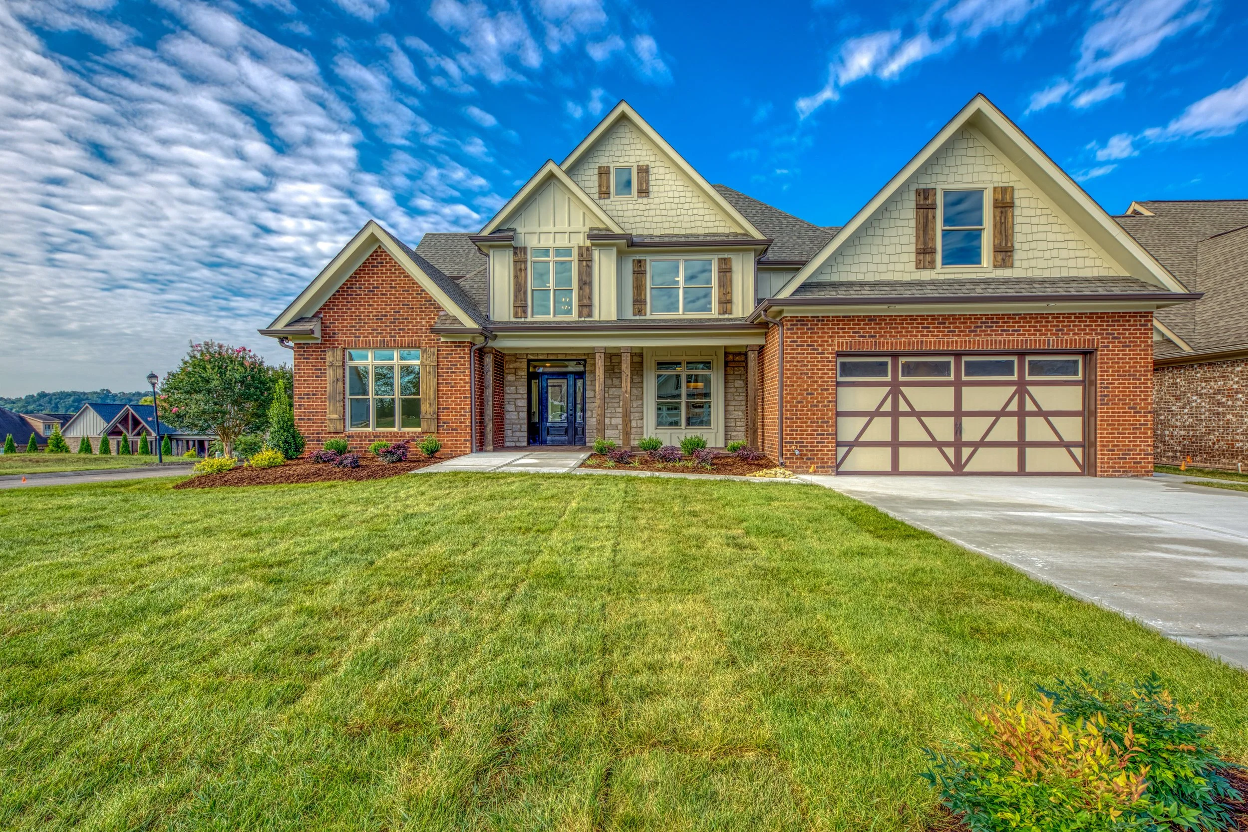A large suburban house with brick and beige siding, multiple gabled roofs, and a two-car garage, surrounded by a well-maintained lawn and landscaped garden, under a partly cloudy blue sky.