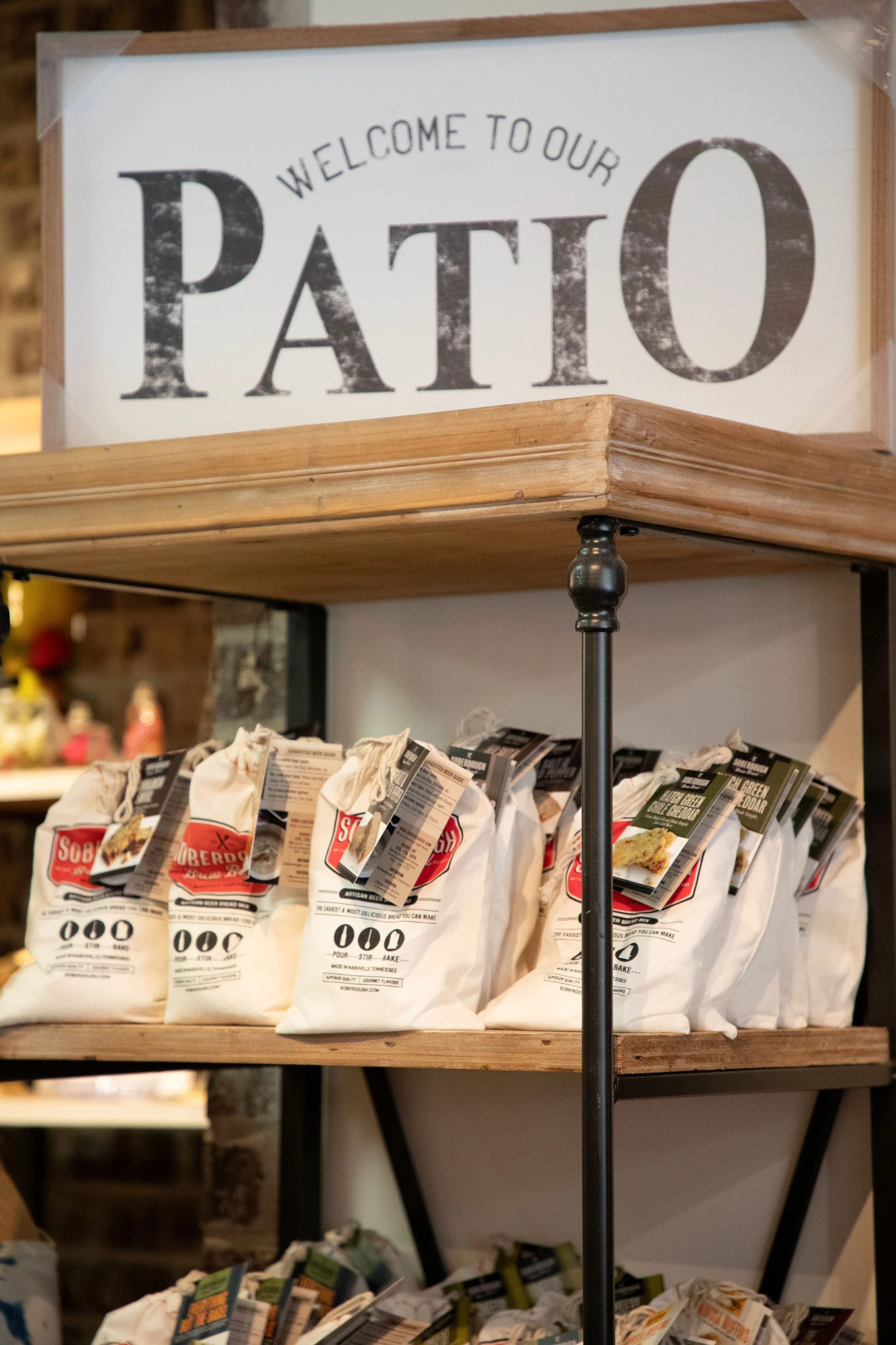 Shelf with bags of Sobe Dough pizza dough mix and other baking ingredients, with a wooden sign that reads 'Welcome to Our Patio'.