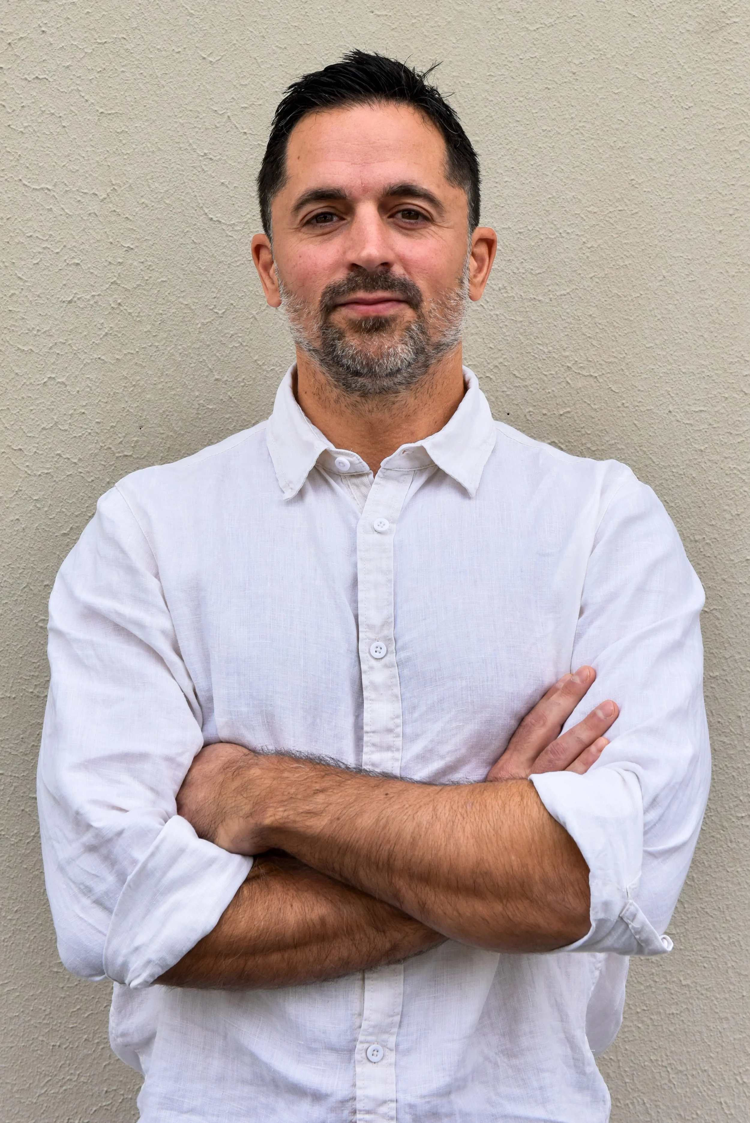 A man with dark hair and a beard, wearing a white button-up shirt, standing with arms crossed in front of a plain textured wall.