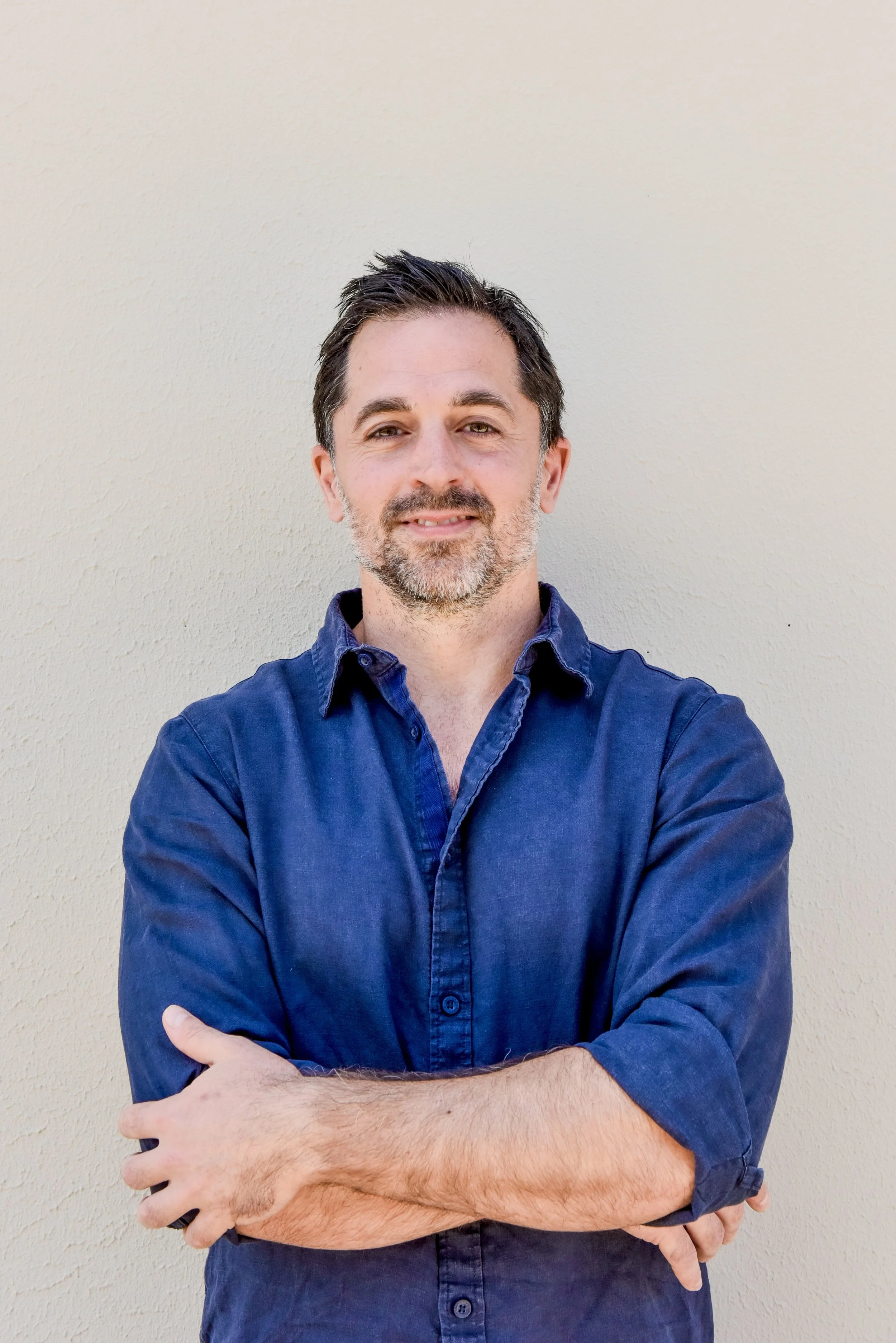 A man with dark hair and a beard, wearing a blue shirt, standing against a plain light-colored wall with arms crossed, smiling slightly.