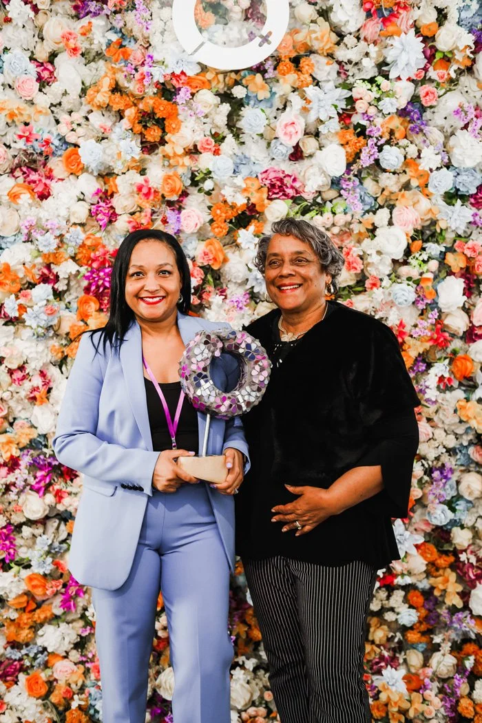 Two women standing in front of a colorful floral backdrop, smiling. The woman on the left holds a circular floral award and a small box. Both are dressed in professional attire.
