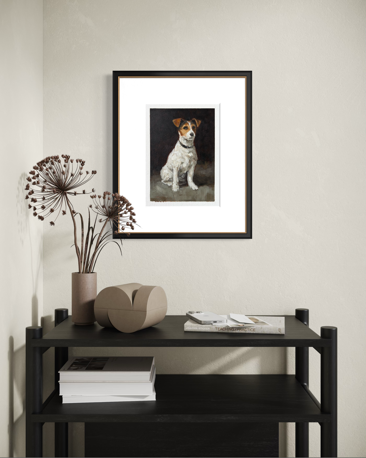 Framed painting of a dog hangs on a beige wall above a black table. The table has a beige abstract sculpture, a tall vase with dried flowers, a stack of white books, and a white remote control. There are additional white books or magazines on a lower