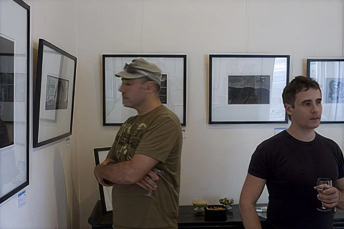 Two men in an art gallery observing framed artwork on the wall.