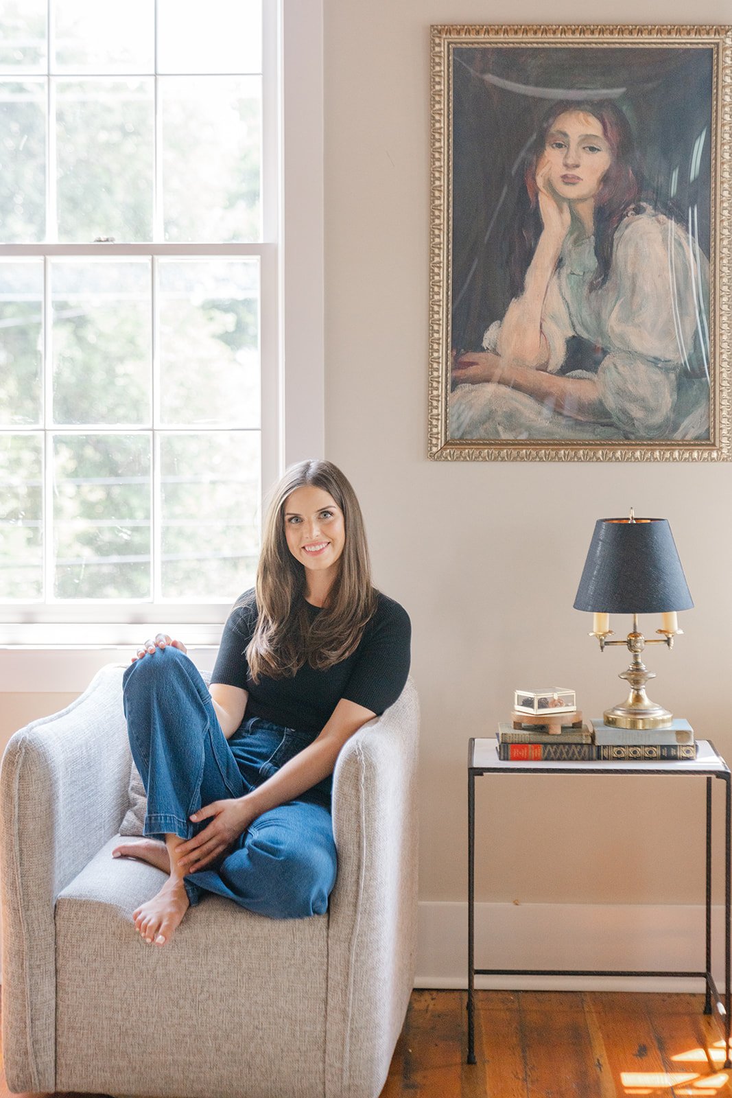 Photograph of woman seated on grey fabric chair indoors, smiling, wearing a black shirt and blue jeans in a bright, airy space with a large art print of a seated woman behind her.