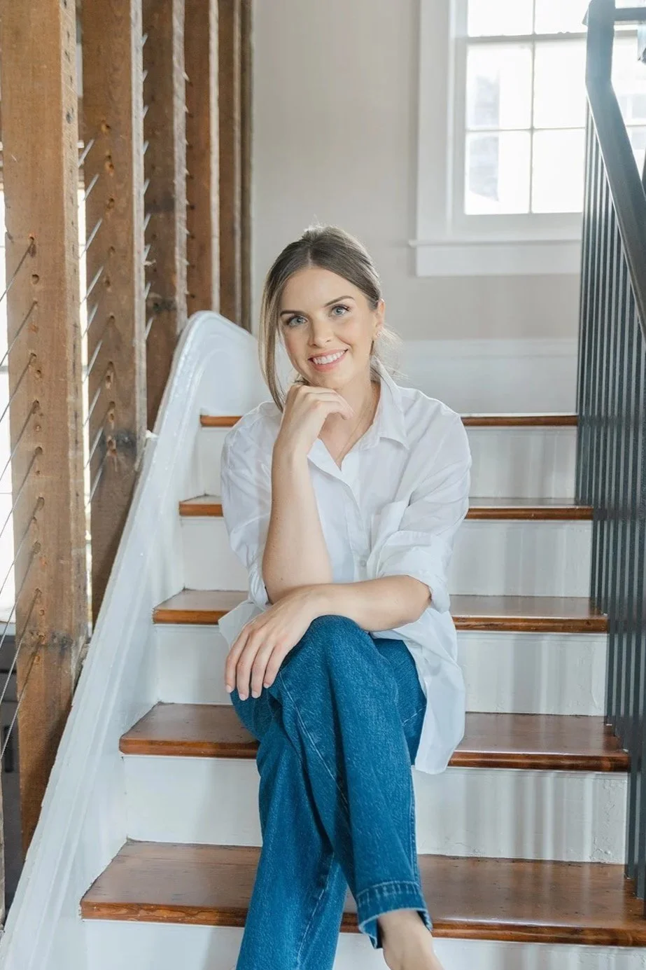 Photograph of woman seated on wooden stairs indoors, smiling, wearing a white button-down shirt and blue jeans in a bright, airy space.