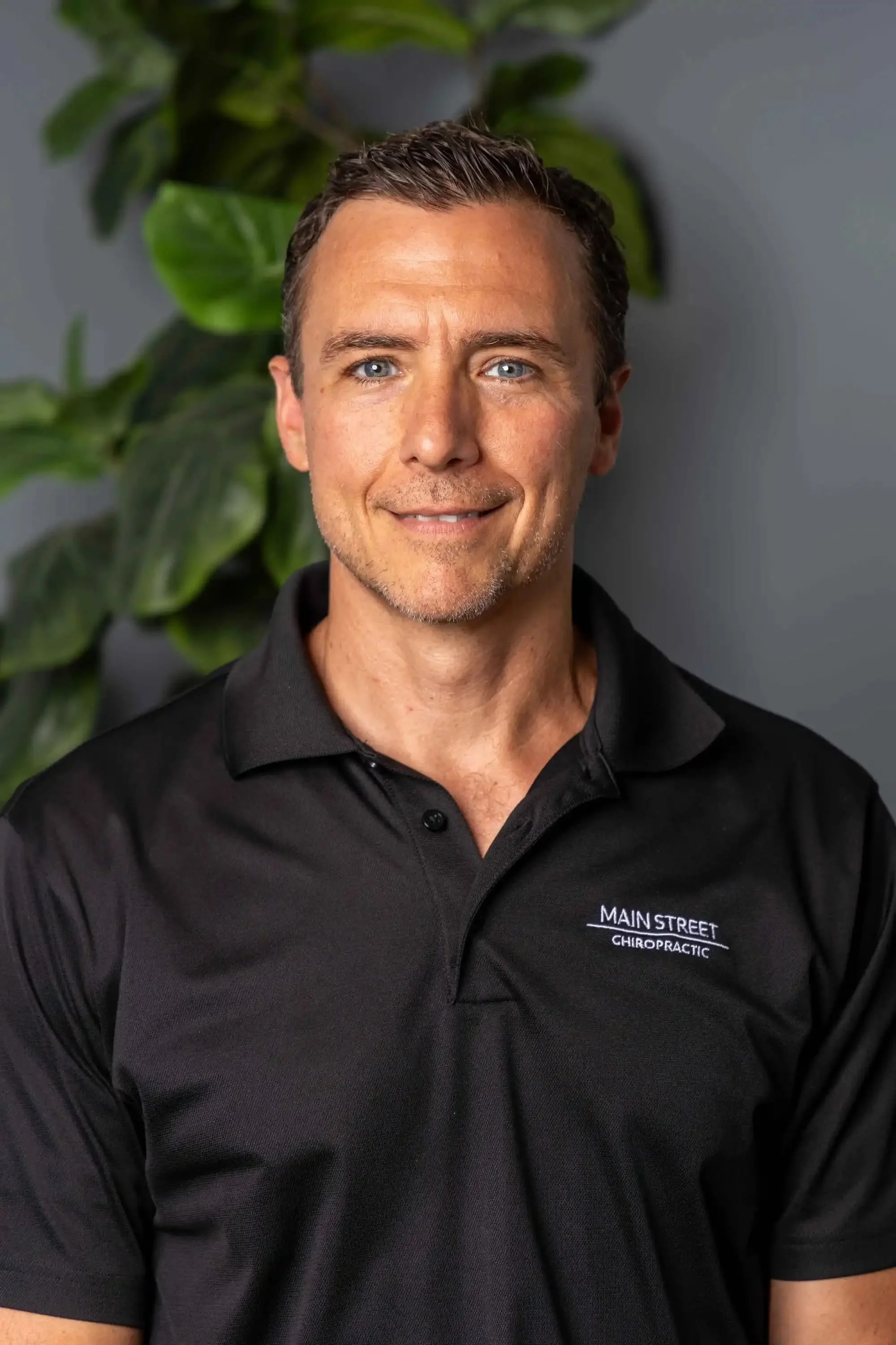 A headshot portrait of Dr. Asa Hadsell wearing a black collared shirt with the "Main Street Chiropractic" logo on the left chest side of the shirt.