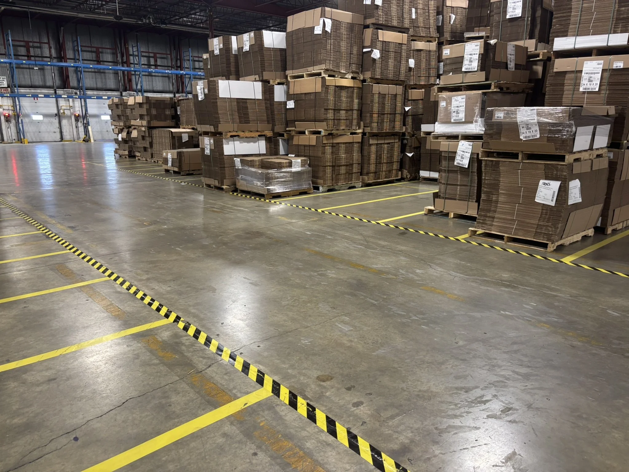 Stacked cardboard boxes on pallets in a warehouse with high shelves and a concrete floor marked with yellow and black safety lines.
