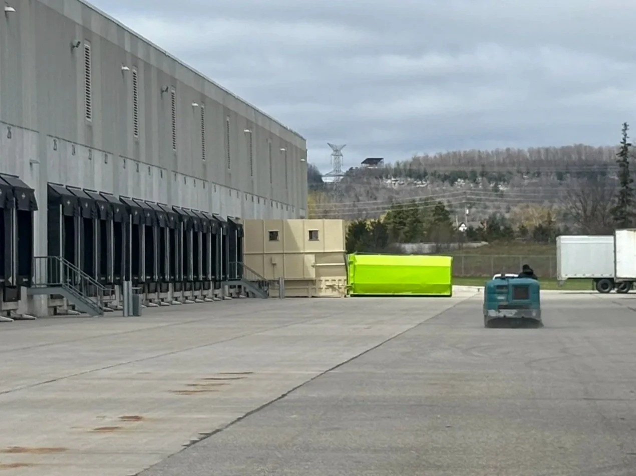 An empty industrial area with a large gray building, a series of black loading dock doors, and a small teal-colored ride-on floor sweeper on a paved lot. Two trucks and some large beige and bright green barriers are visible in the background.