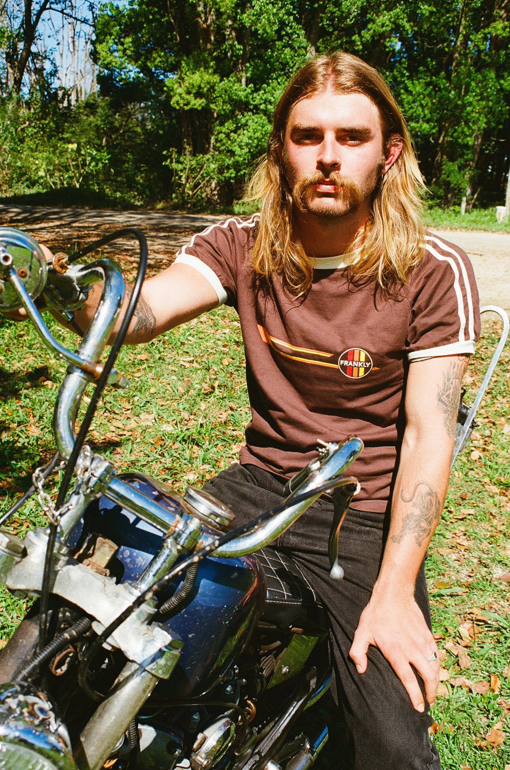 A man with long blonde hair and tattoos on his arms, wearing a brown t-shirt with a 'Frankly' logo, is sitting on a motorcycle outdoors with trees and greenery in the background.