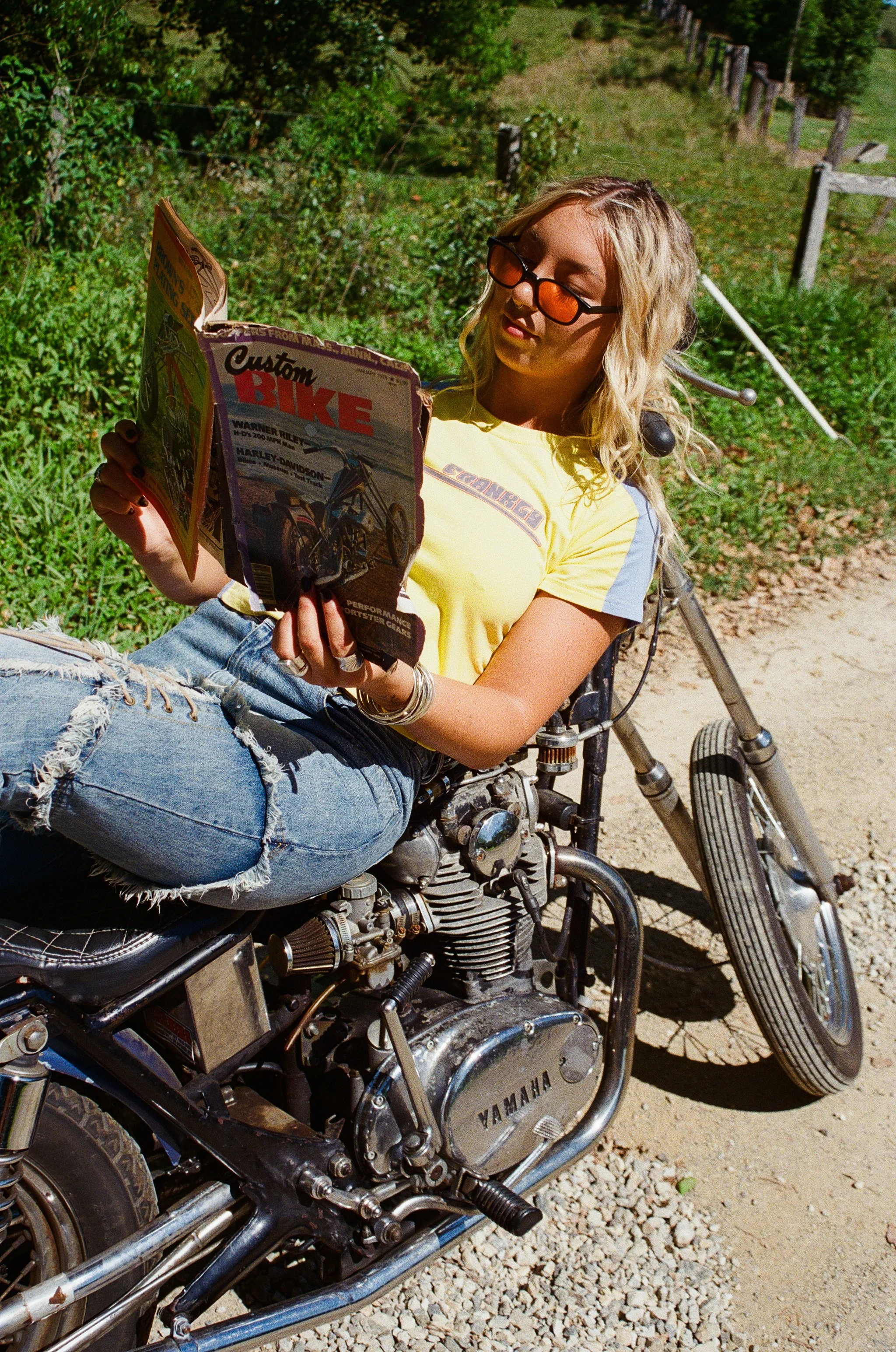 A young woman with wavy blonde hair wearing orange-tinted sunglasses, a yellow T-shirt, and ripped jeans, sitting on a motorcycle while reading a magazine outdoors on a dirt path with greenery and fencing in the background.