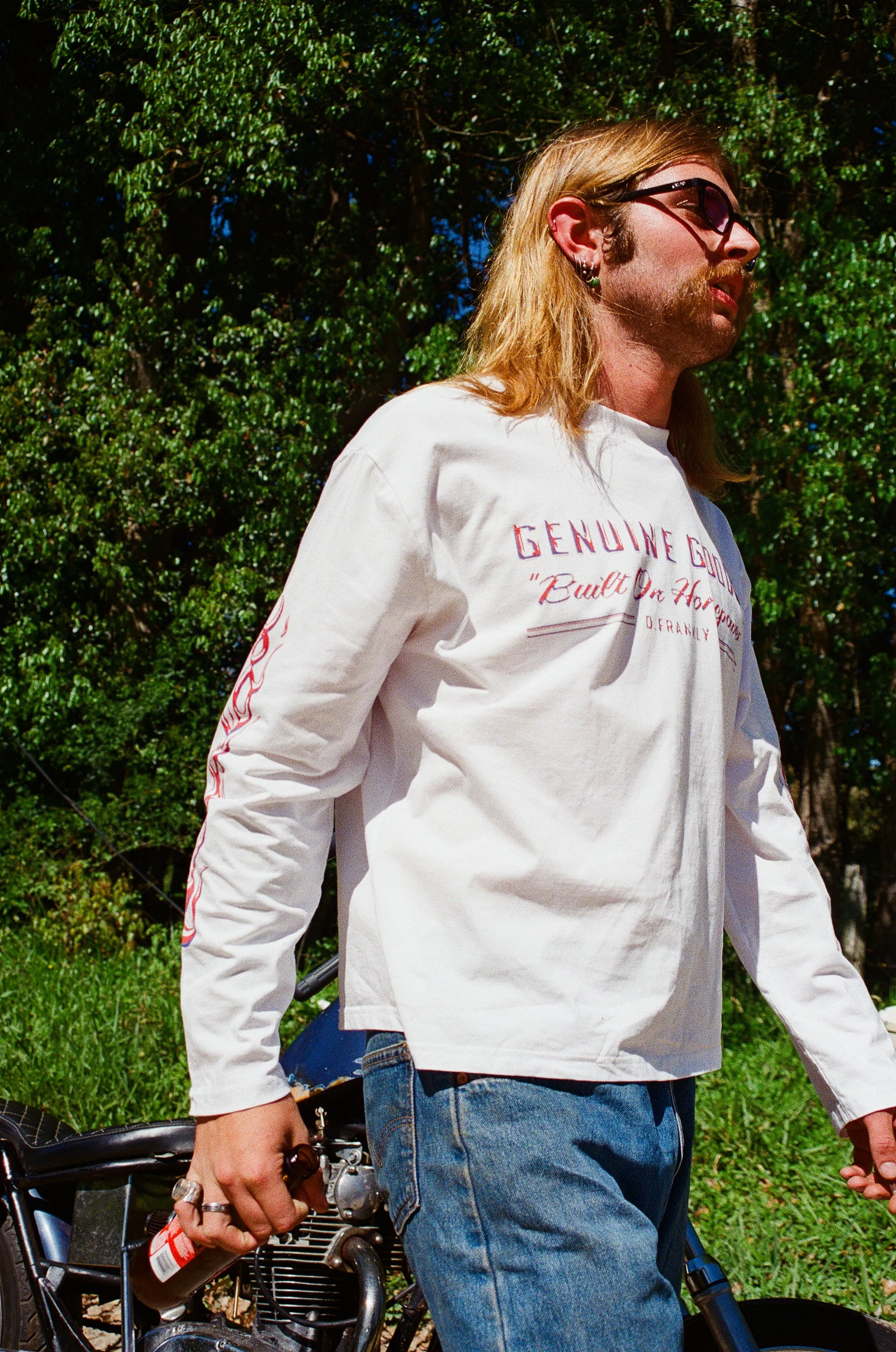 A person with long blonde hair, wearing sunglasses and a white long-sleeve shirt, standing outdoors next to a bicycle with greenery and trees in the background.