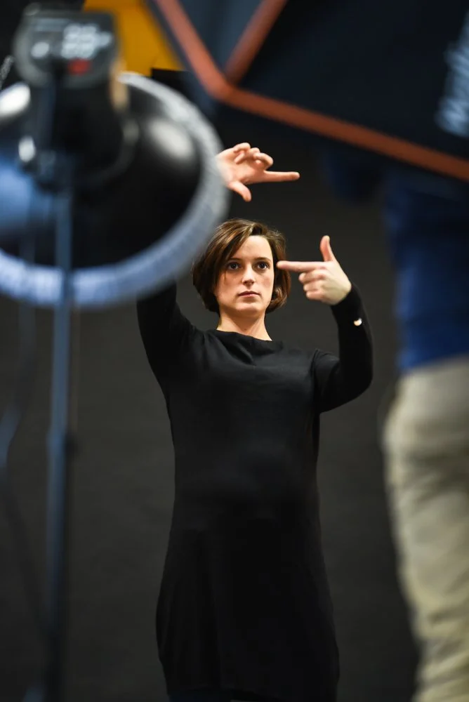 A woman with short brown hair and wearing a black long-sleeve shirt is standing indoors, gesturing with her hands, with a focused expression on her face.