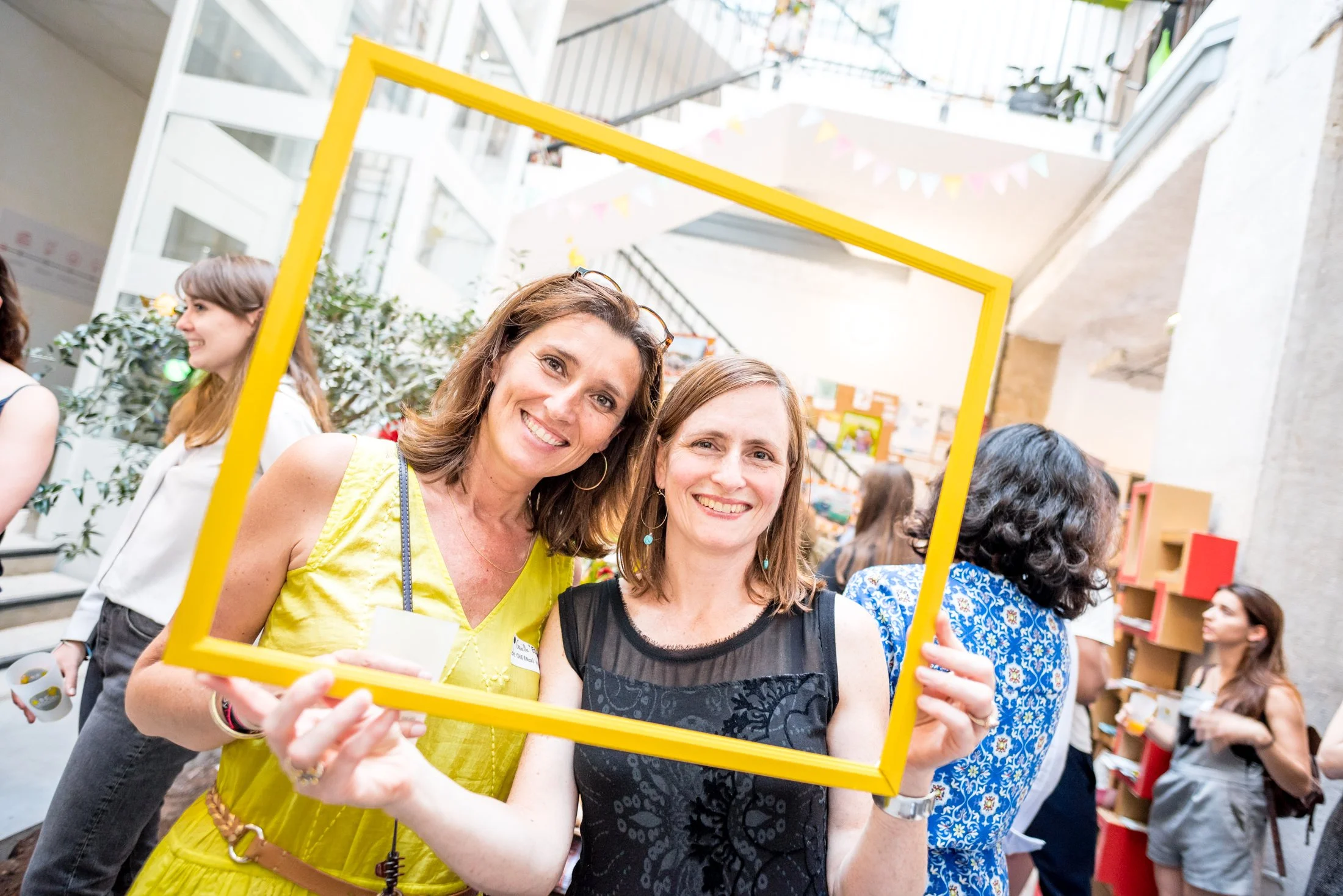 Two women smiling and holding a yellow picture frame at a party or event, with other people in the background at an indoor venue decorated with bunting.