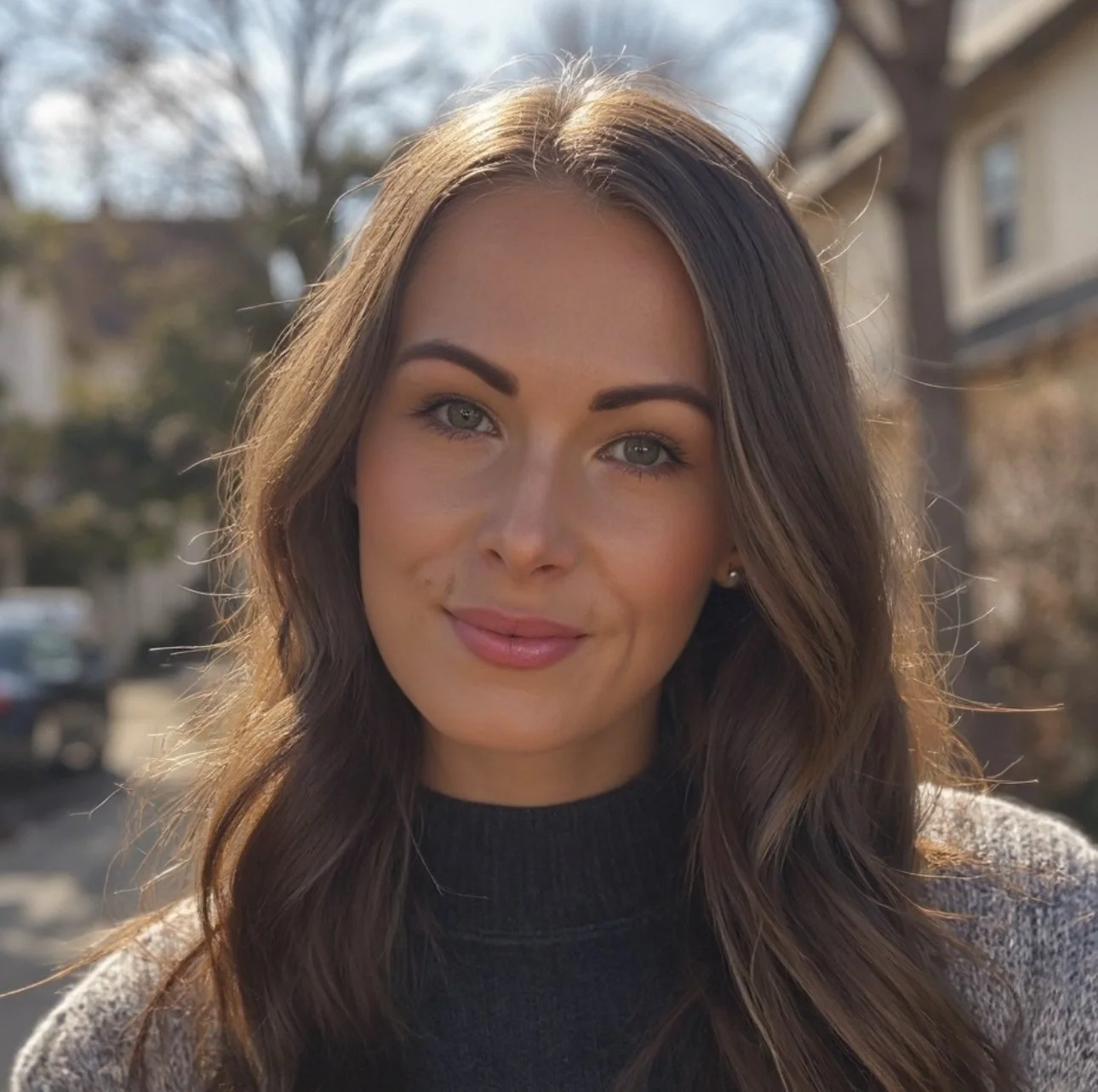 A young woman with long brown hair and blue eyes smiling outdoors on a sunny day, with blurred trees and houses in the background.