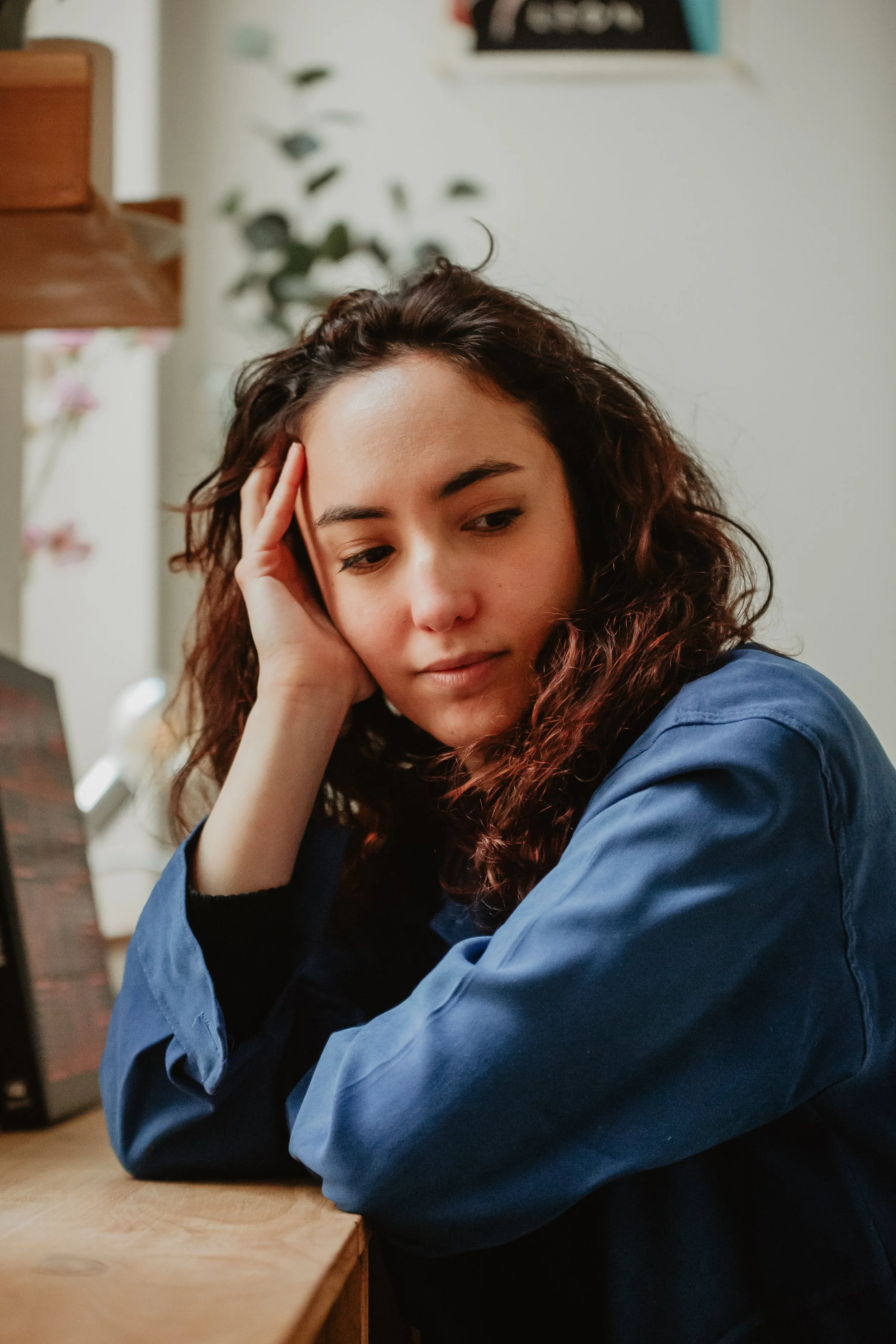 Une jeune femme aux cheveux bruns frisés, portant un pull noir et une veste bleue, repose sa tête sur sa main, assise à une table en bois.