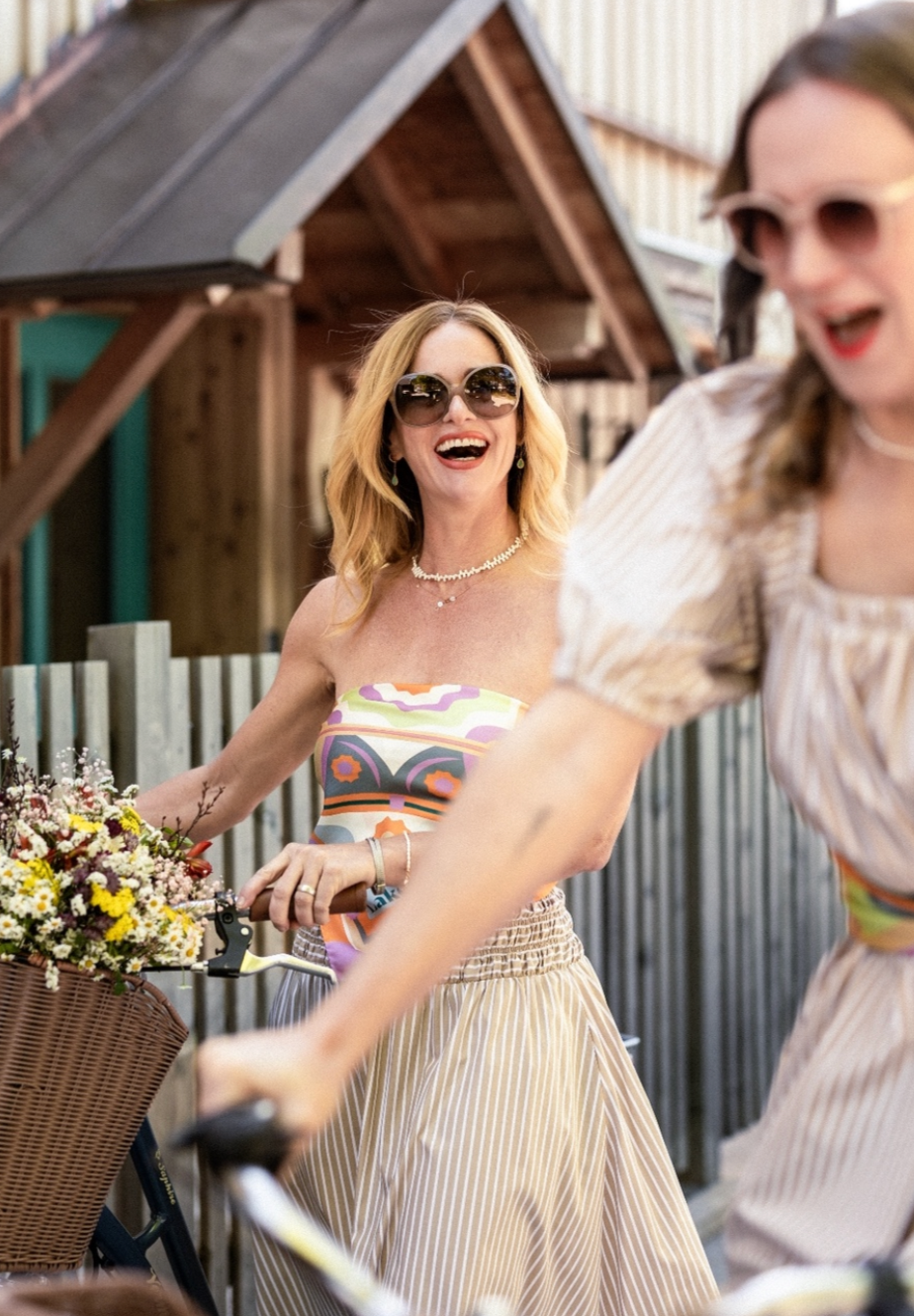 Zwei junge Frauen radfahren in einem Garten, eine Frau lacht und trägt eine bunte Tops und eine Streifenhose, die andere Frau führt das Fahrrad an.