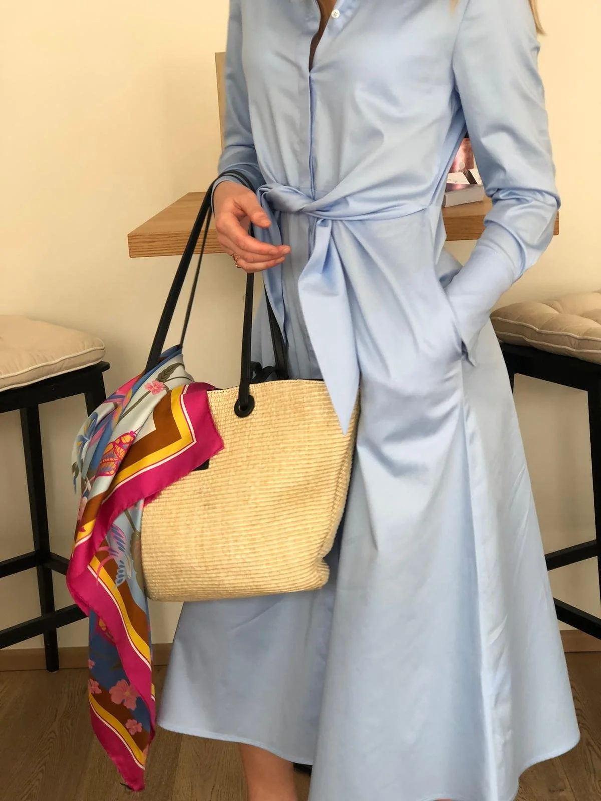 Eine Frau trägt ein hellblaues Kleid mit langen Ärmeln und einer Krawatte, die gebunden ist. Sie hält eine große strohhütenartige Tasche mit schwarzen Griffen, die mit einem bunten Schal geschmückt ist, vor ihrem Körper.