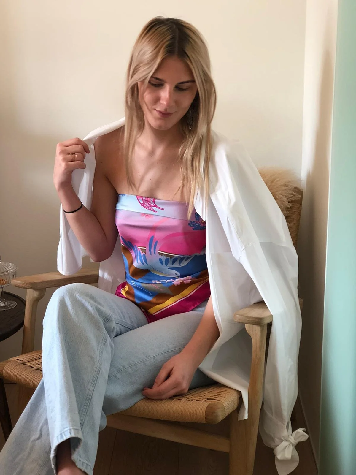 Eine junge Frau sitzt auf einem Stuhl, trägt ein buntes, schulterfreies Kleid mit einem blauen, pinken und weißen Muster, und eine weiße Jacke. Sie hat lange, blonde Haare und schaut nach unten.